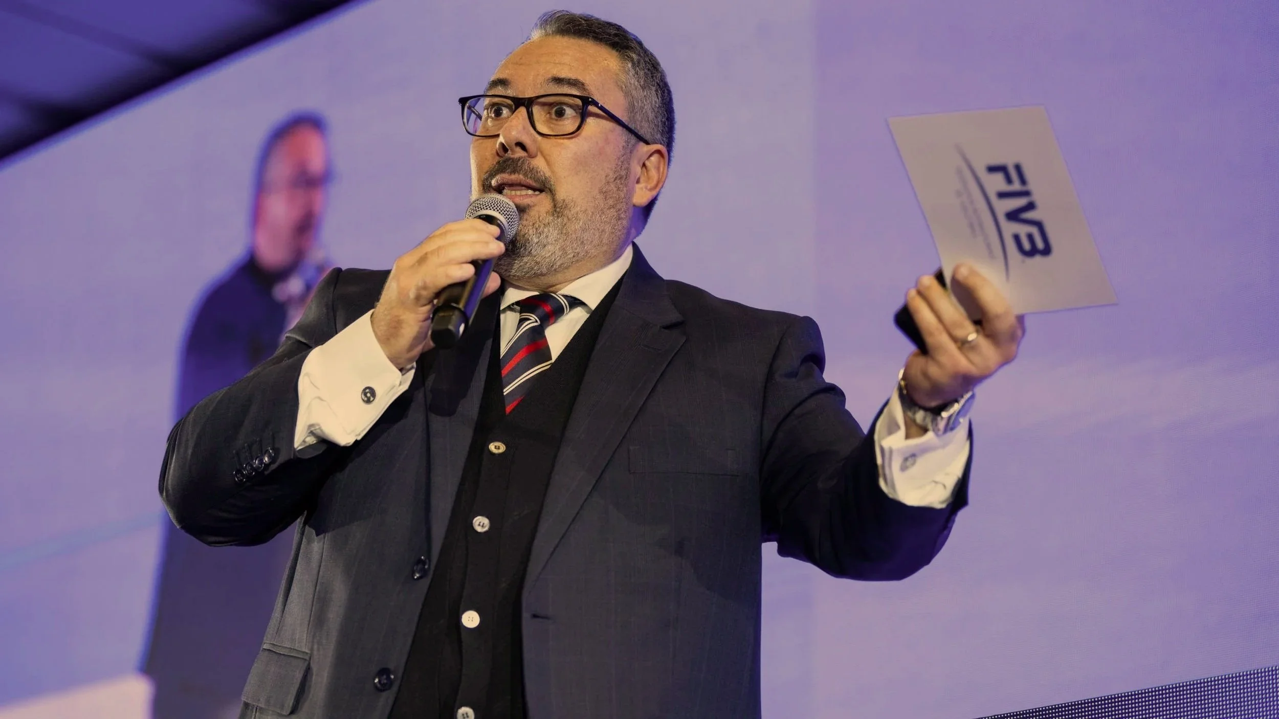 Hombre de negocios con gafas y barba, vestido con traje oscuro y corbata a rayas, habla en un micrófono en un escenario, sosteniendo un papel con letras que dicen 'FIB'.