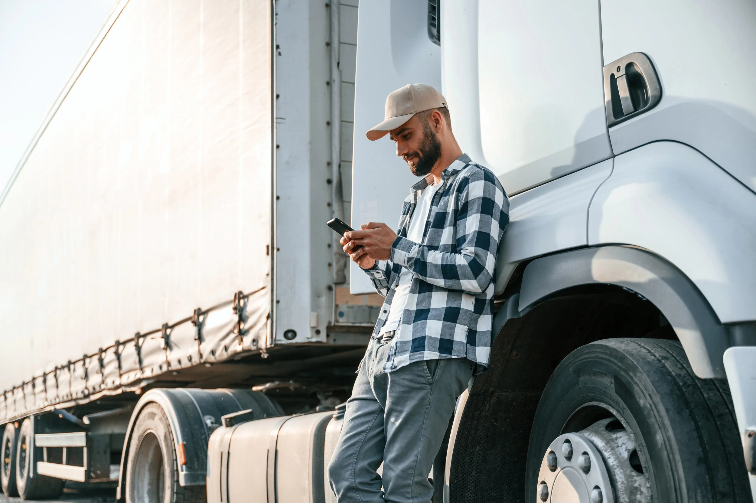 A man with a beard wearing a beige cap and a blue and white checkered shirt standing next to a large white semi-truck trailer, looking at his cellphone.