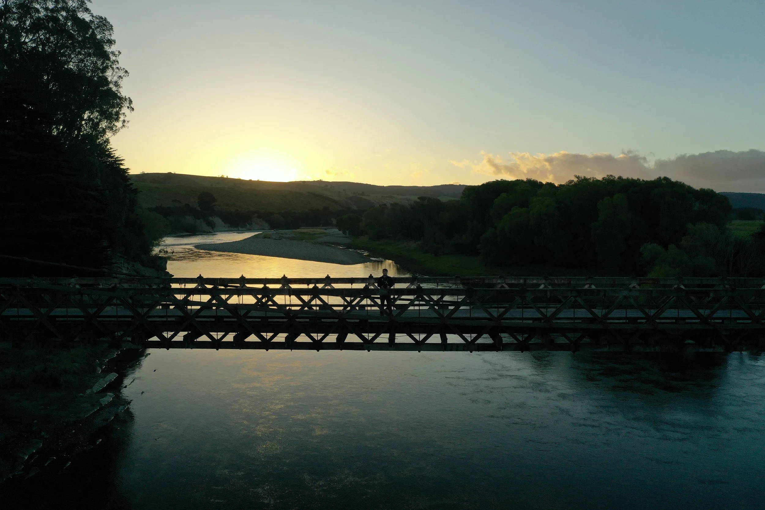 Sonnenuntergang über einem Fluss mit einer Brücke und Bäumen an den Ufern, eine Person steht auf der Brücke.