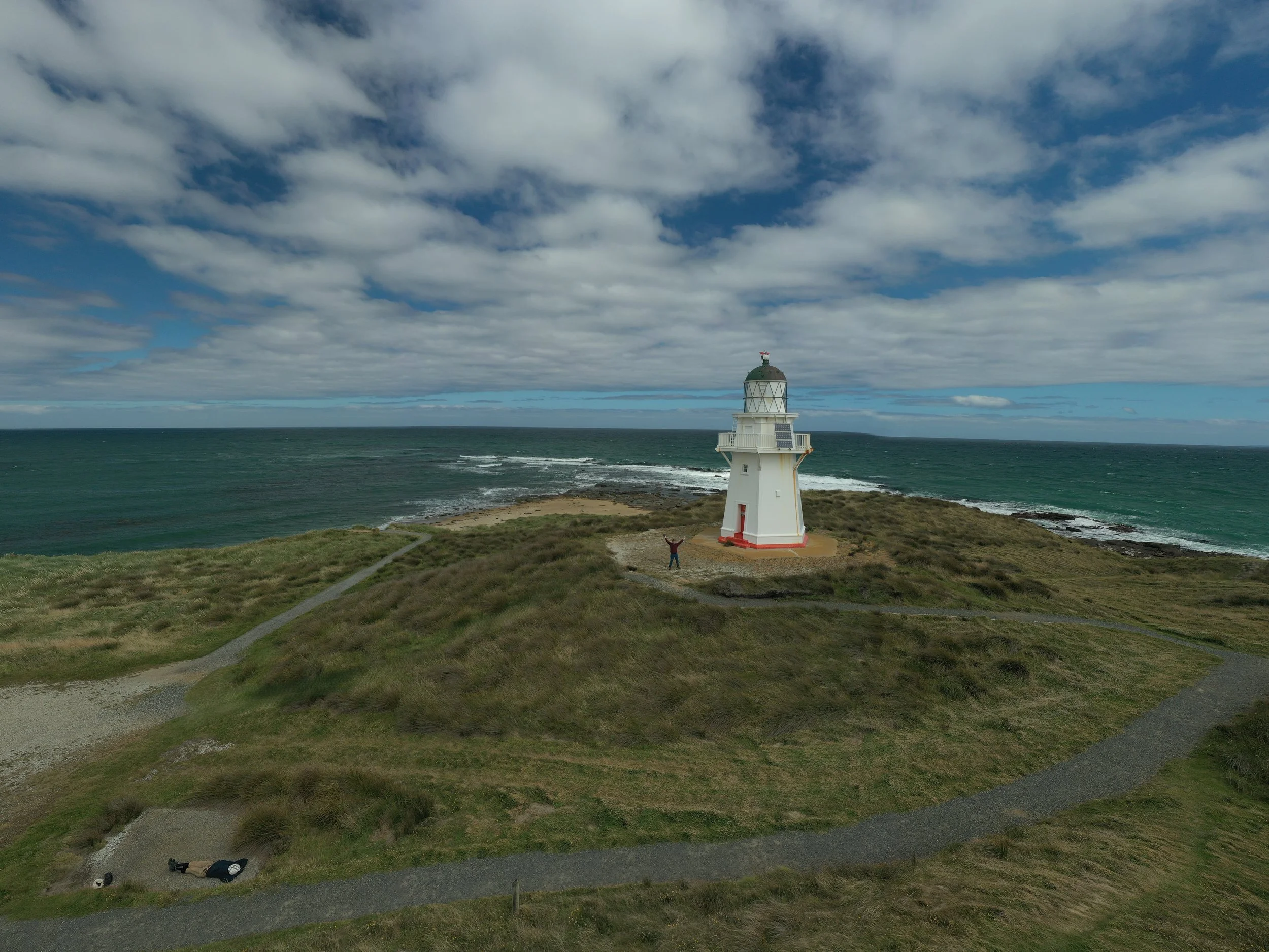 Leuchtturm an der Küste mit Himmel und Wolken, umgeben von grünem Gras und Wegen, eine Person mit erhobenen Armen steht vor dem Leuchtturm.