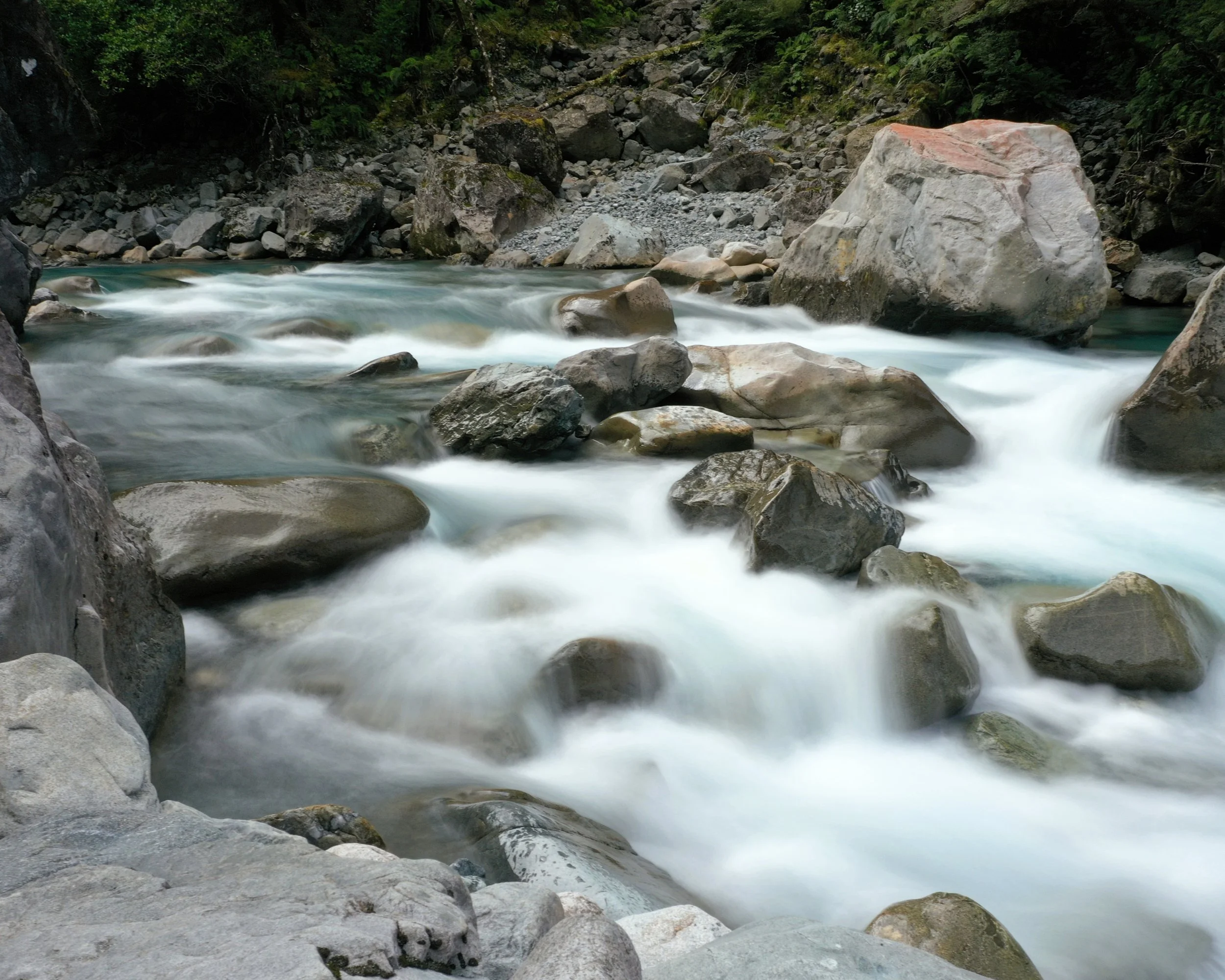 Ein plätschernder Fluss mit großen und kleinen Steinen in einer natürlichen Umgebung mit Bäumen und Felsen.