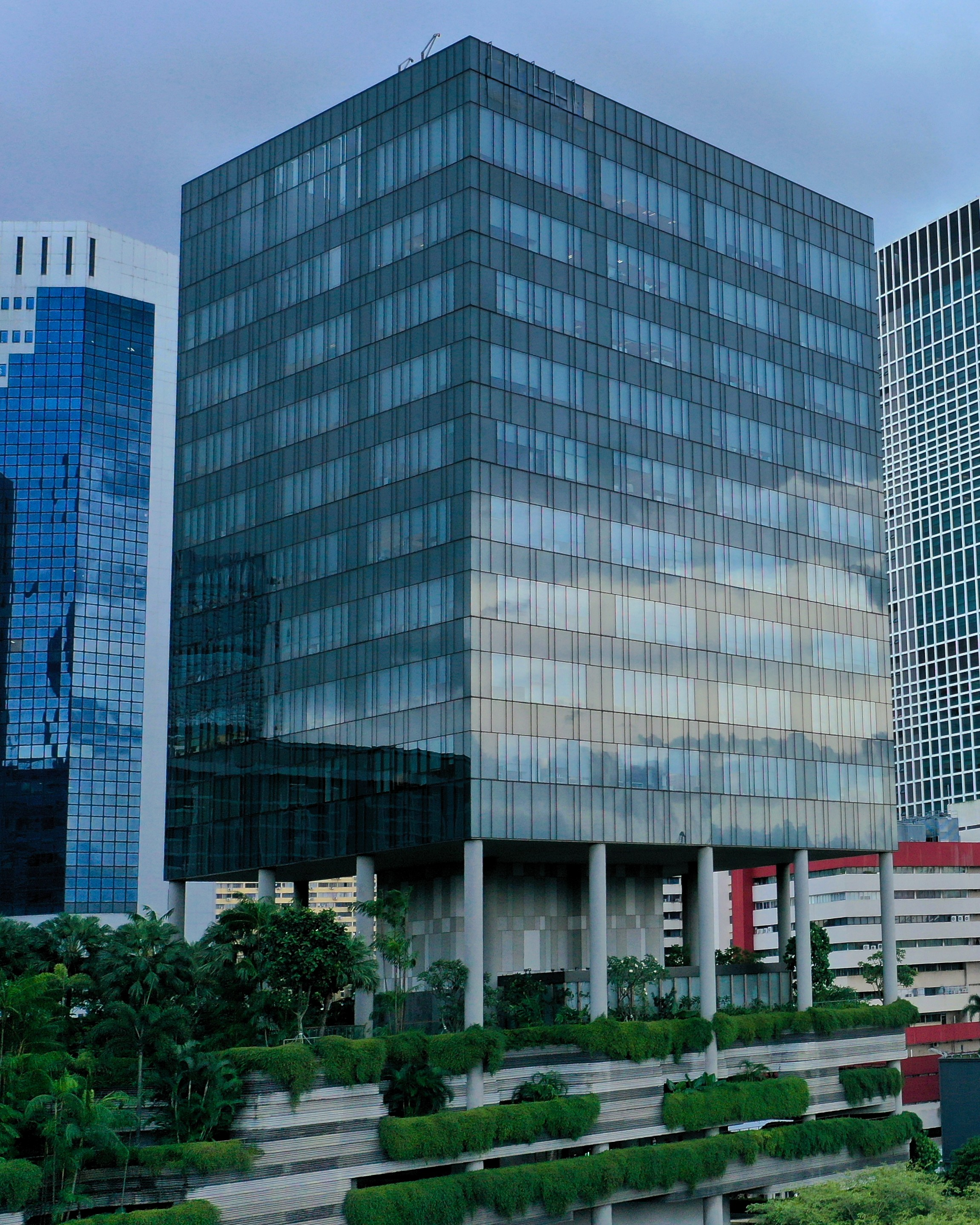 Ein modernes Hochhaus mit reflektierendem Glasfassade, umgeben von anderen Wolkenkratzern und begrünten Terrassen.