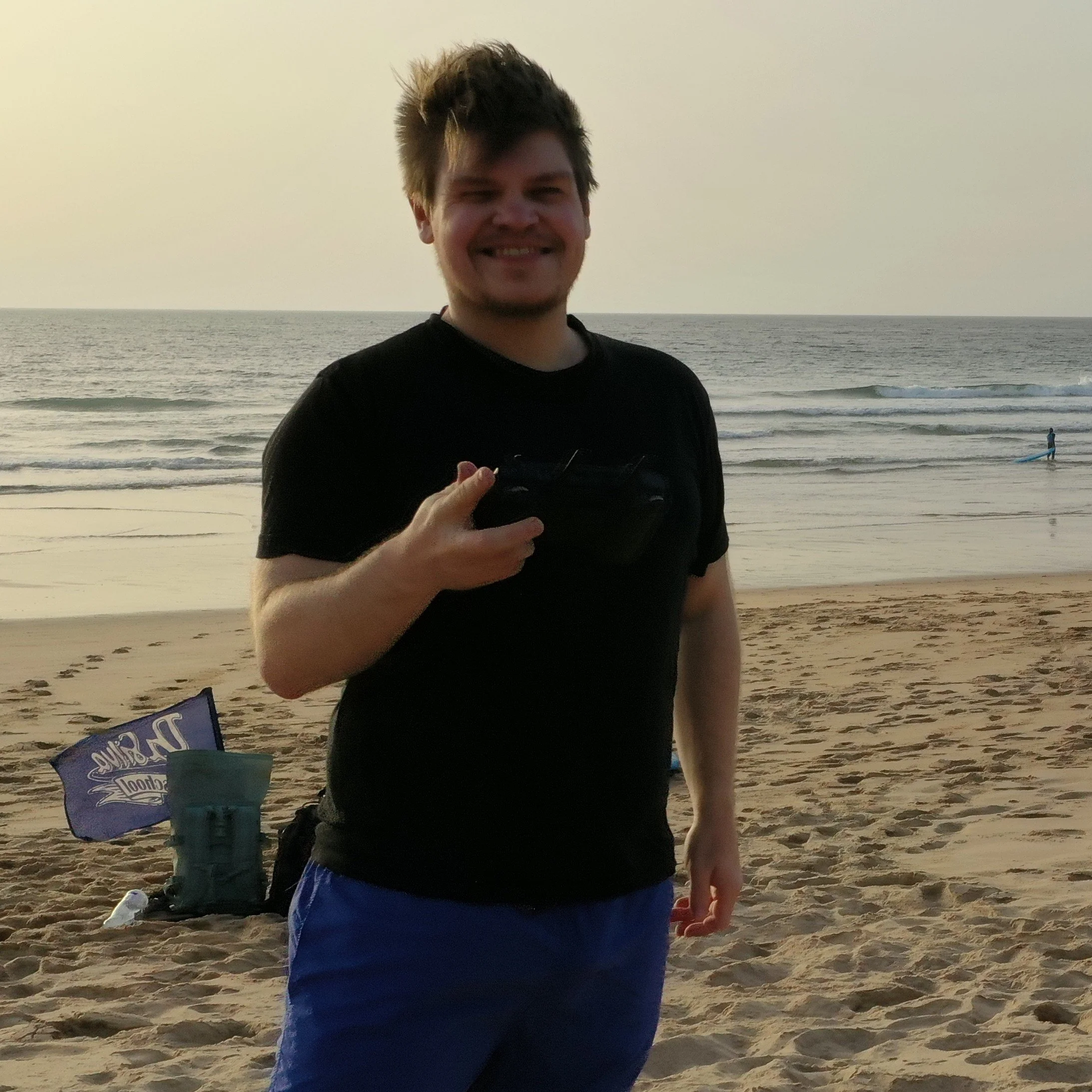 Ein junger Mann am Strand, hält eine Fernbedienung und lächelt, im Hintergrund das Meer und eine Person beim Surfen.