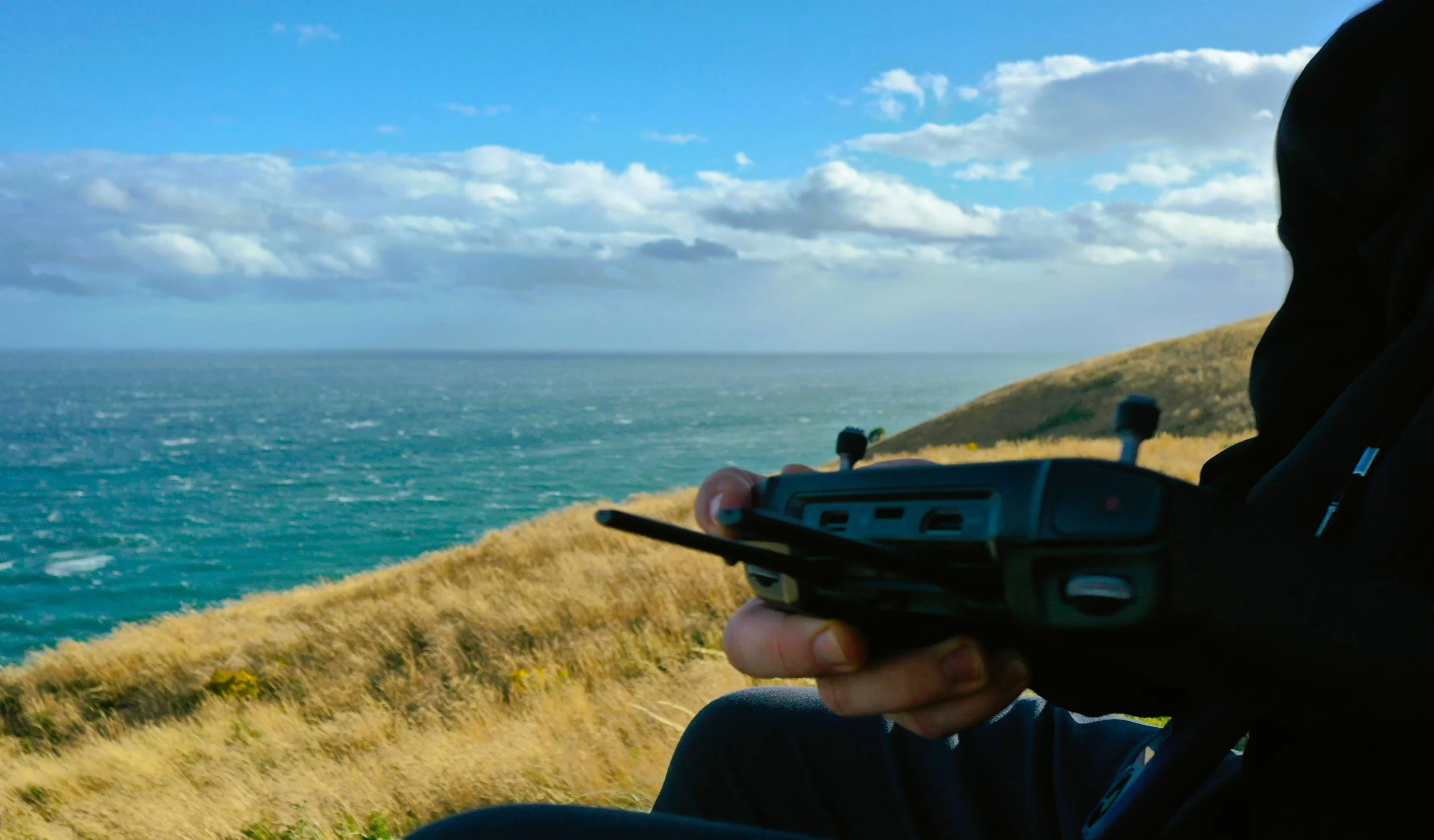 Person hält eine Fernbedienung in der Hand, Blick auf die Küste und das Meer bei sonnigem Himmel.