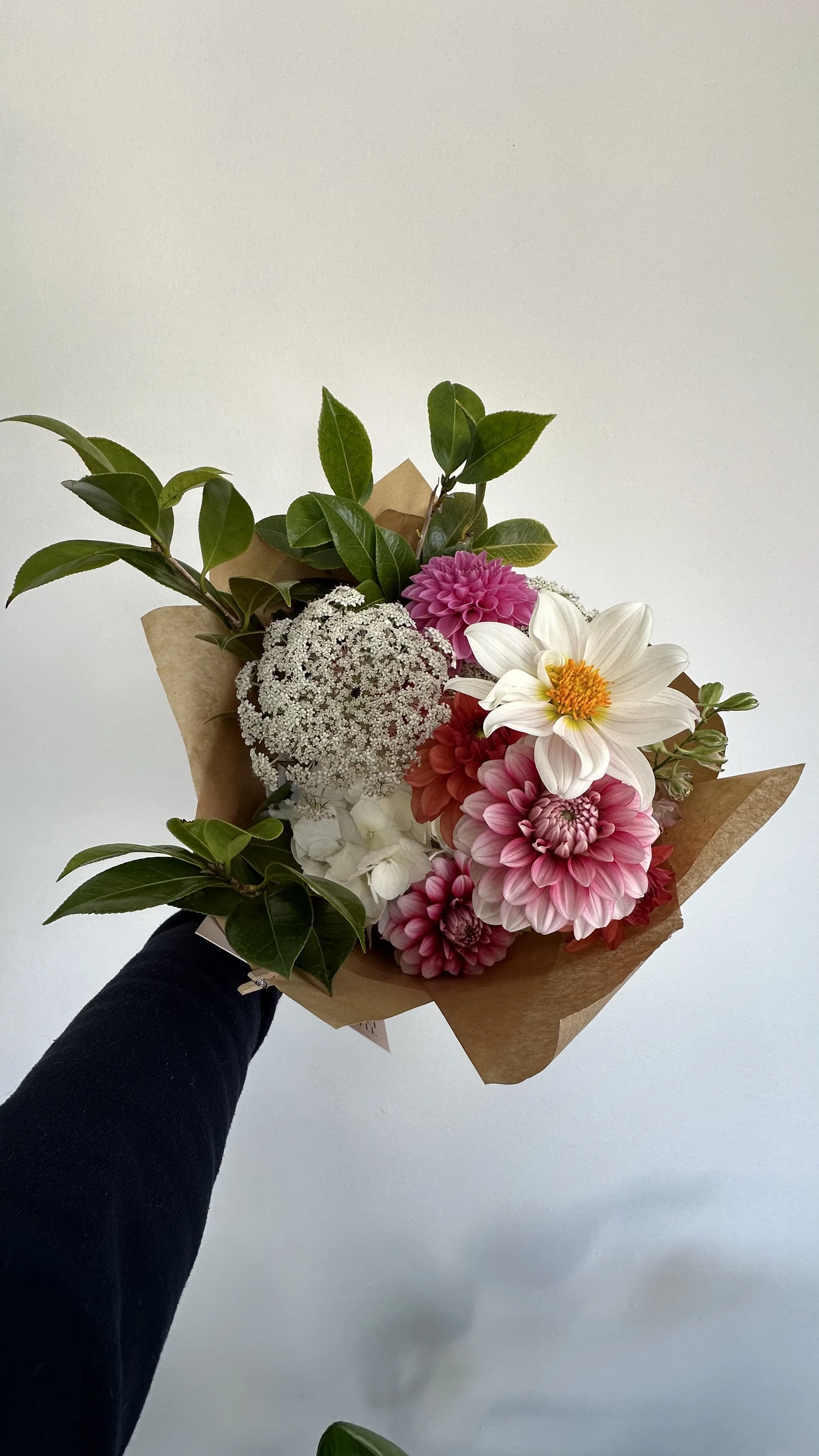 Colorful flower bouquet with roses, dahlias, and other flowers wrapped in brown paper, held by a person wearing a black long-sleeve shirt against a plain light background.