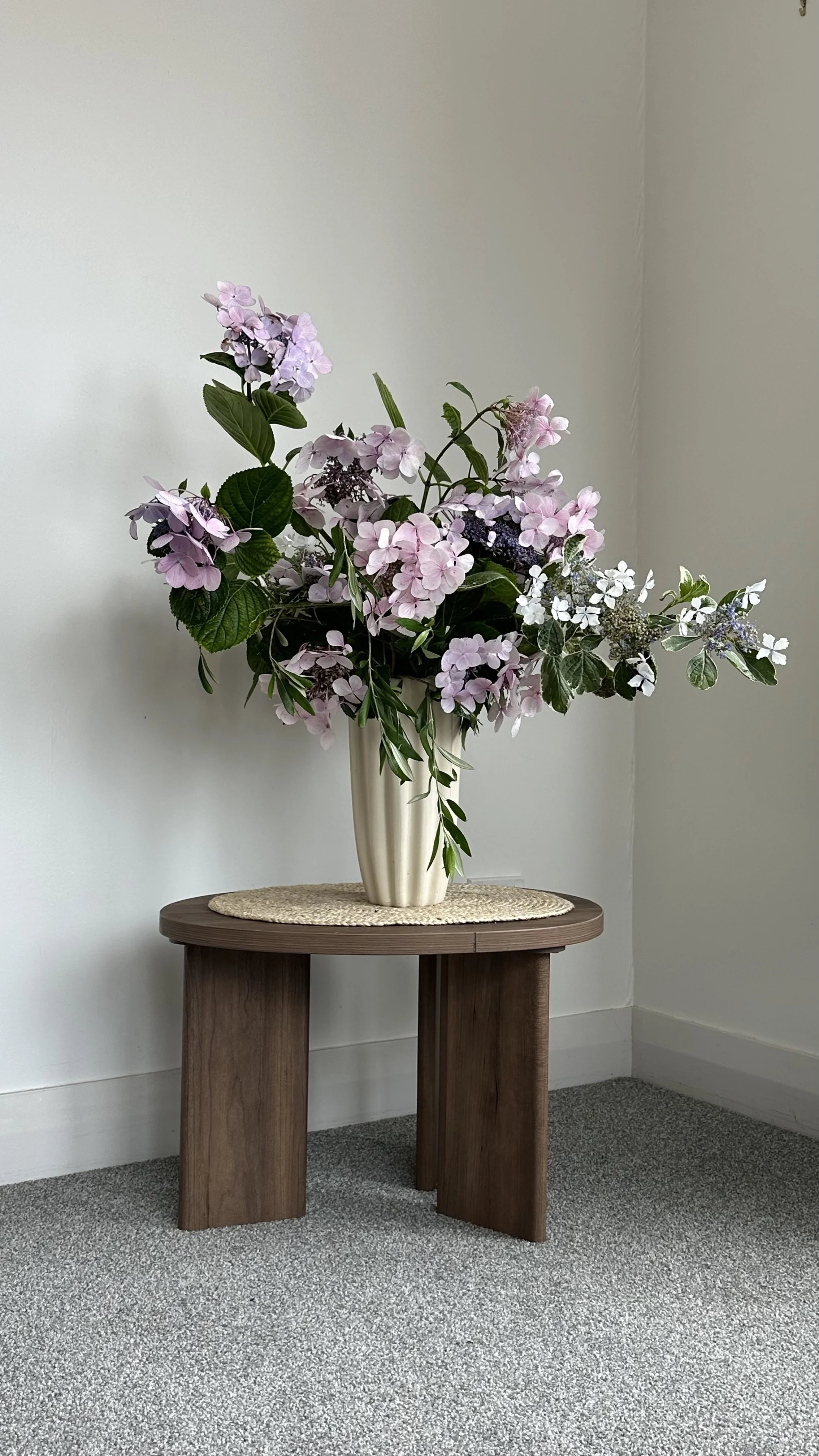 A large bouquet of pink and white hydrangea flowers in a white ceramic vase on a small wooden table against a white wall.