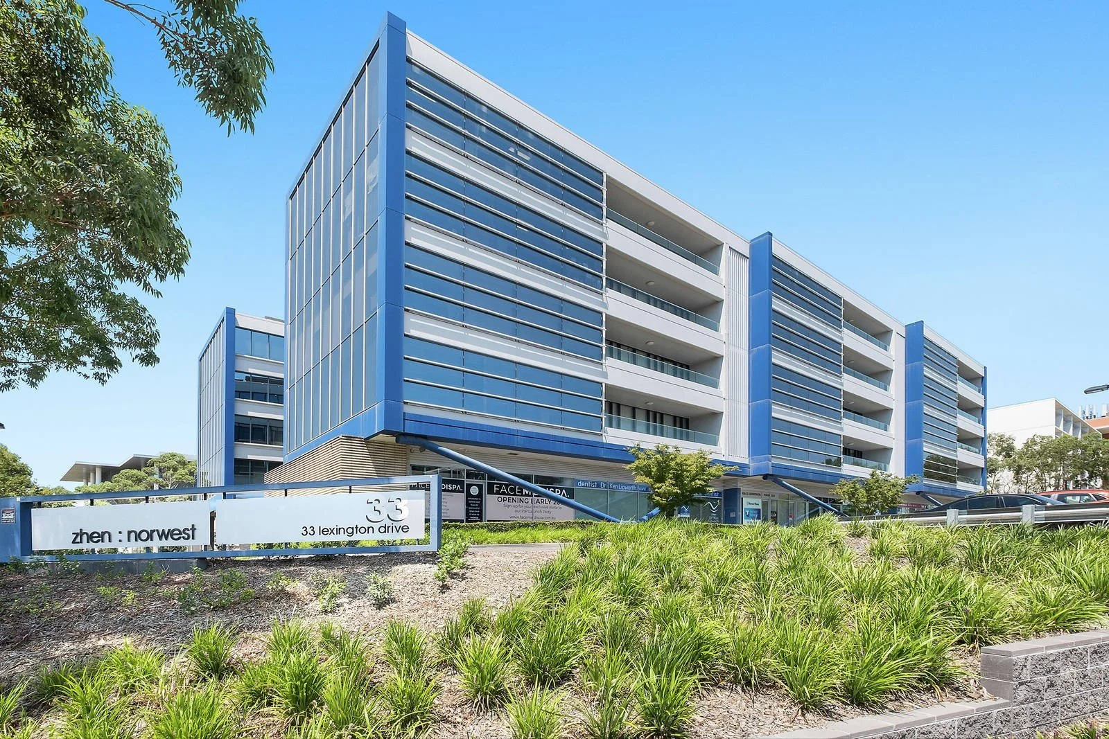 Modern multi-story commercial building with blue and white exterior, large glass windows, and balconies, situated in a landscaped area with green bushes and trees under a clear blue sky.