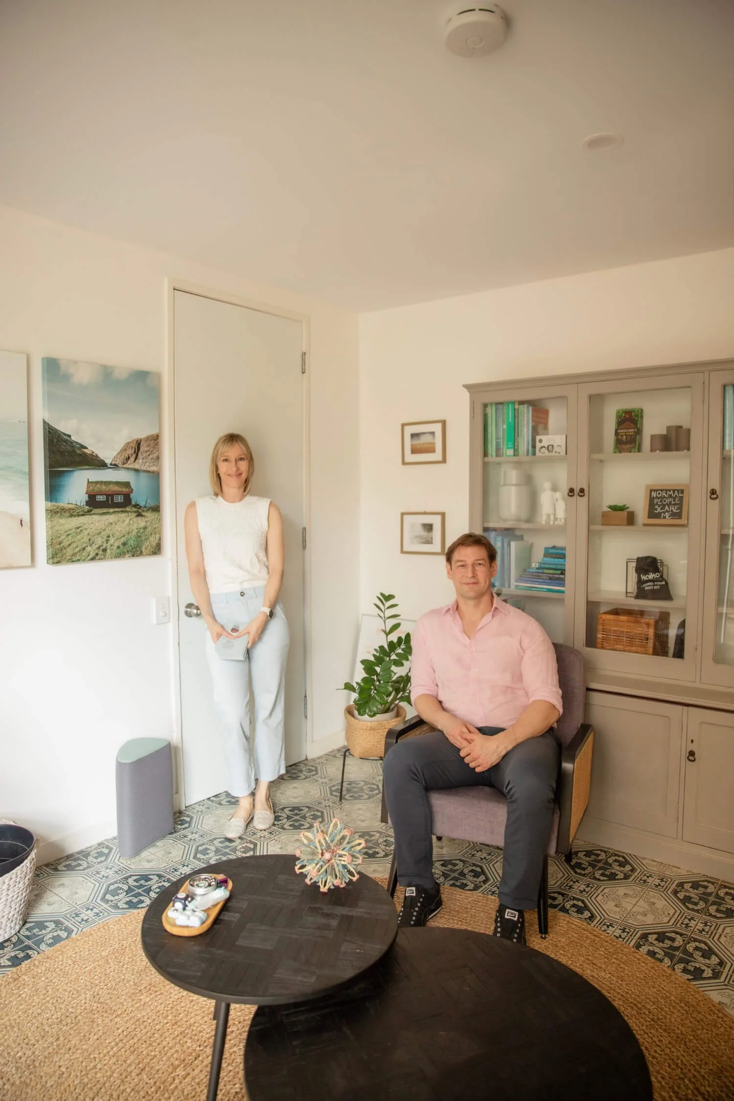 A woman with blonde hair, wearing a white sleeveless top and light jeans, standing near a closed door in a living room. A man with brown hair, wearing a pink shirt and dark pants, sitting in a purple armchair. The room has patterned floor tiles, a round woven rug, a black coffee table, a bookshelf filled with books and decorative items, and a potted plant.