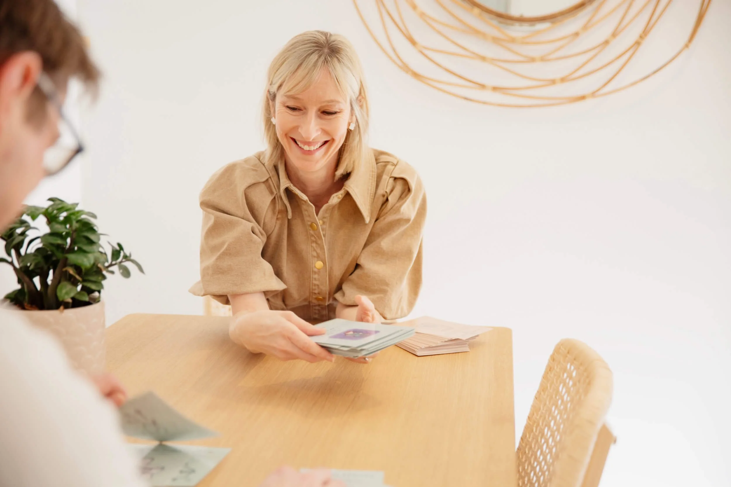 Smiling woman playing a card game with a person whose face is not visible, sitting at a wooden table with a potted plant, light-colored walls, and a decorative wall art.