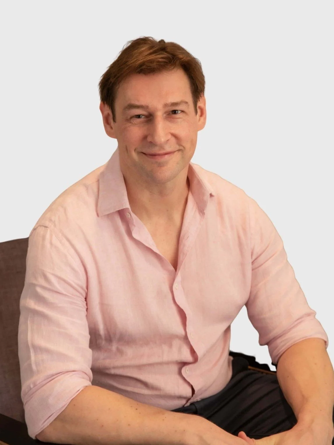 A man with light skin and brown hair smiling, wearing a light pink dress shirt with sleeves rolled up, sitting on a chair against a plain background.