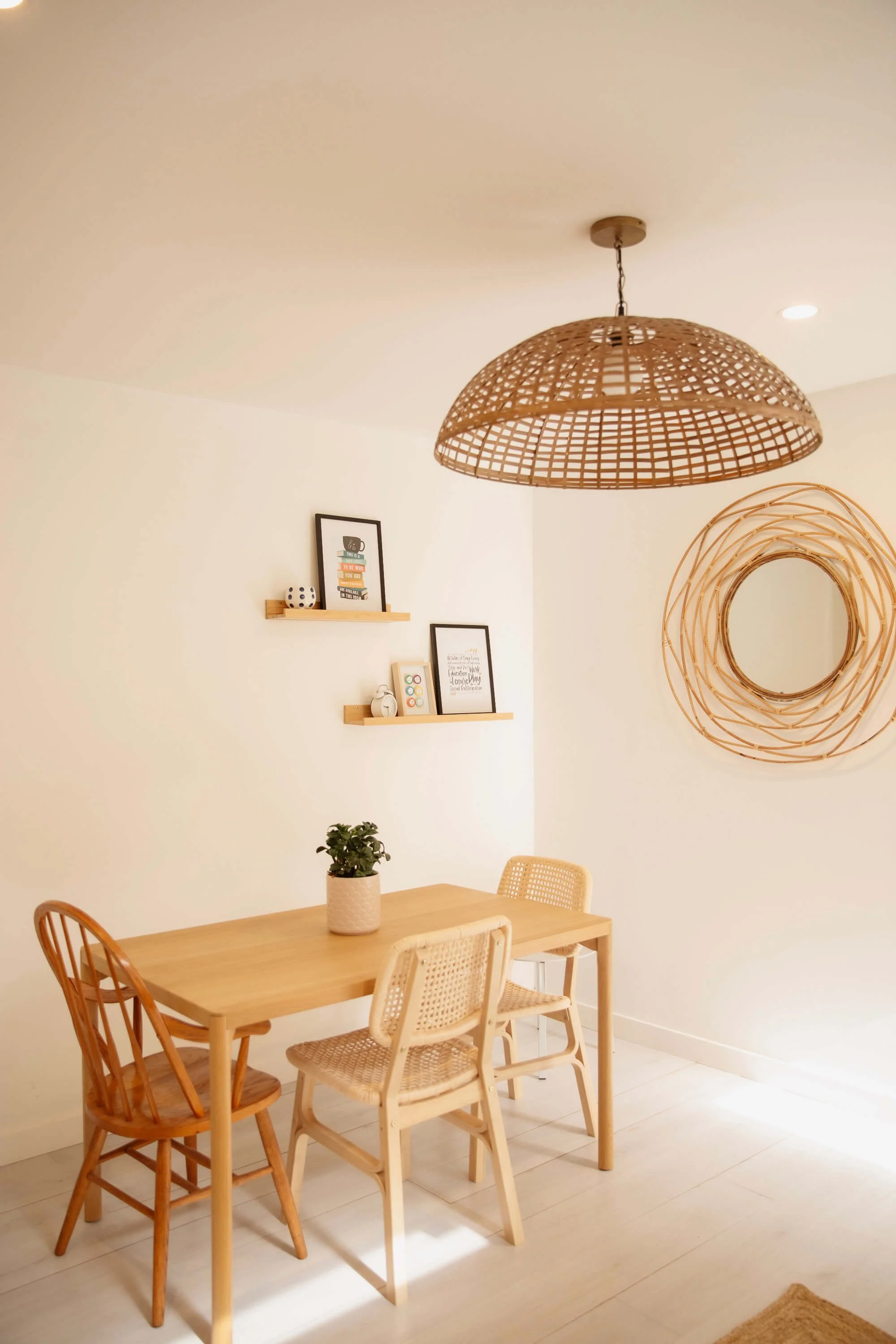 Minimalist dining area with a wooden table, three different chairs, potted plant, decorative wall mirror, wall art, and a woven ceiling light in neutral tones.