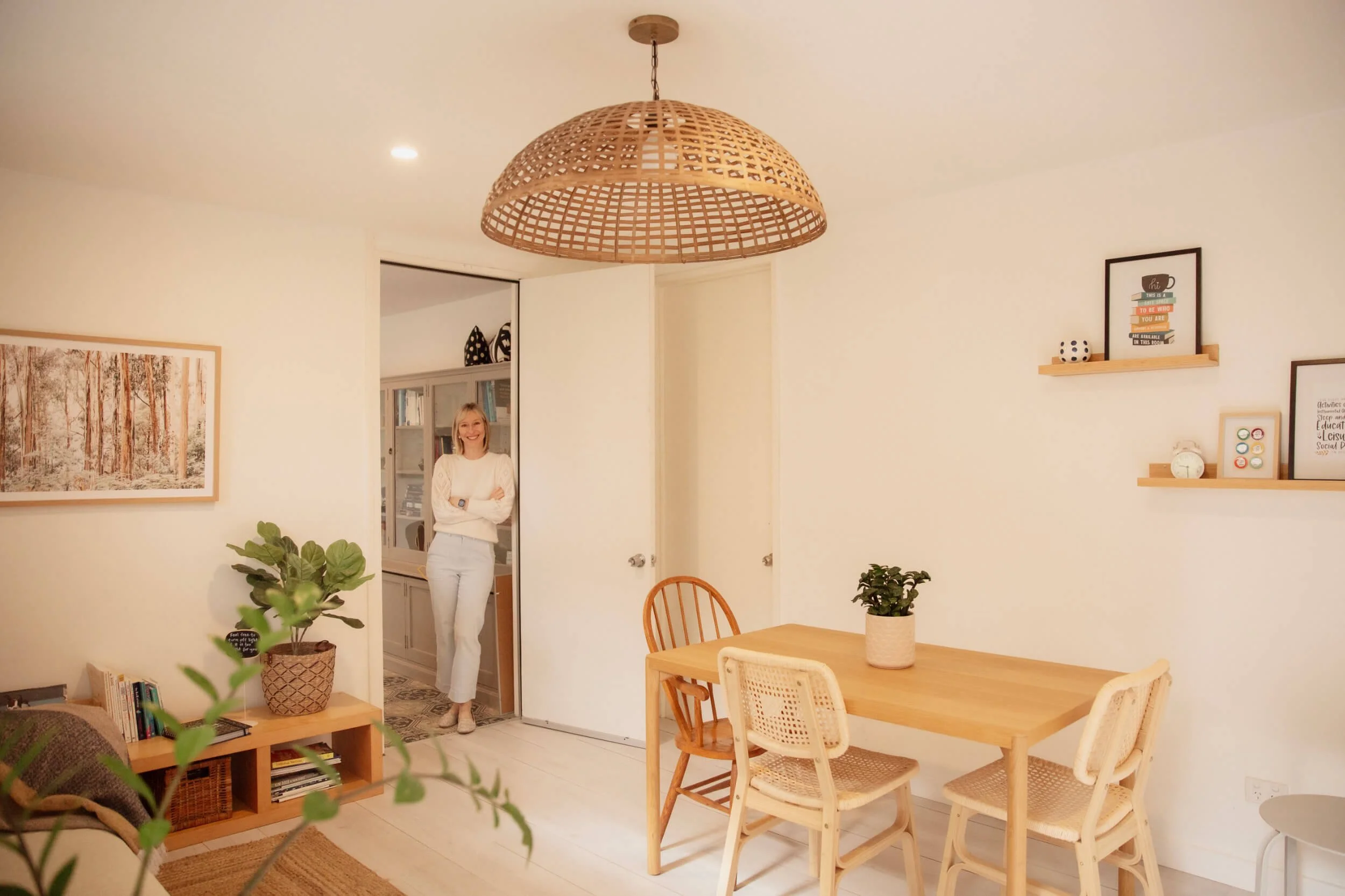 Occupational therapist Natalie Evans standing in the doorway of a brightly lit living room, smiling, with a dining table and chairs, potted plants, and framed pictures on the walls.
