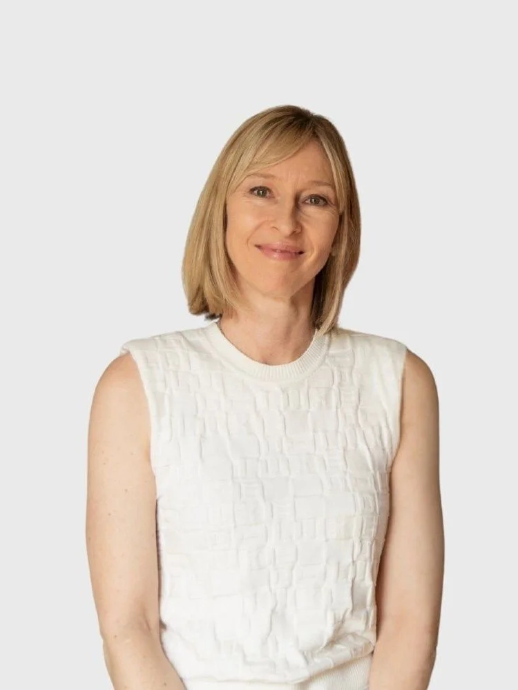 A woman with shoulder-length blonde hair, wearing a sleeveless white textured top, smiling against a plain white background.