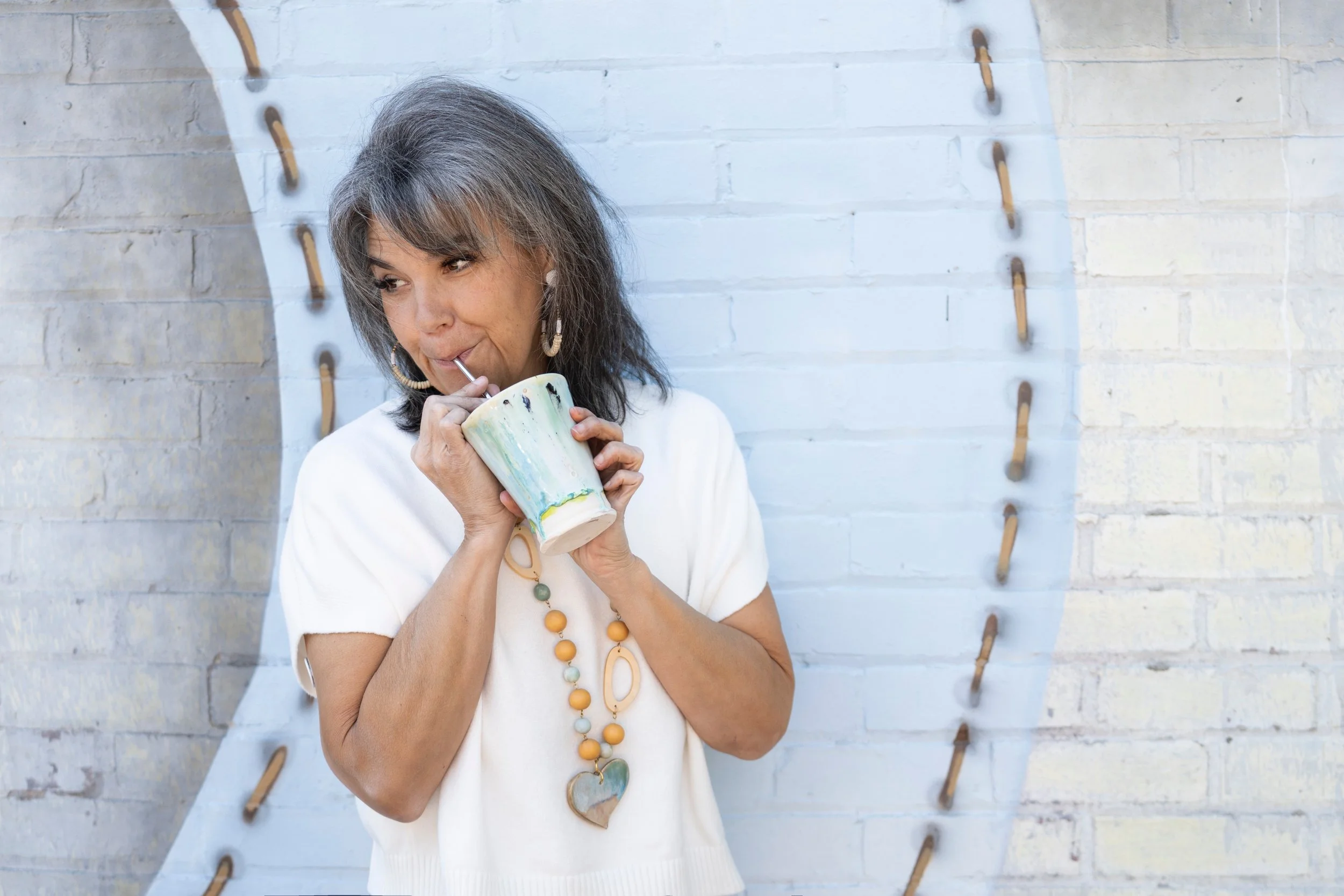 A middle-aged woman with gray hair drinking from a ceramic mug with a straw, standing outdoors against a light-colored brick wall with decorative hooks.