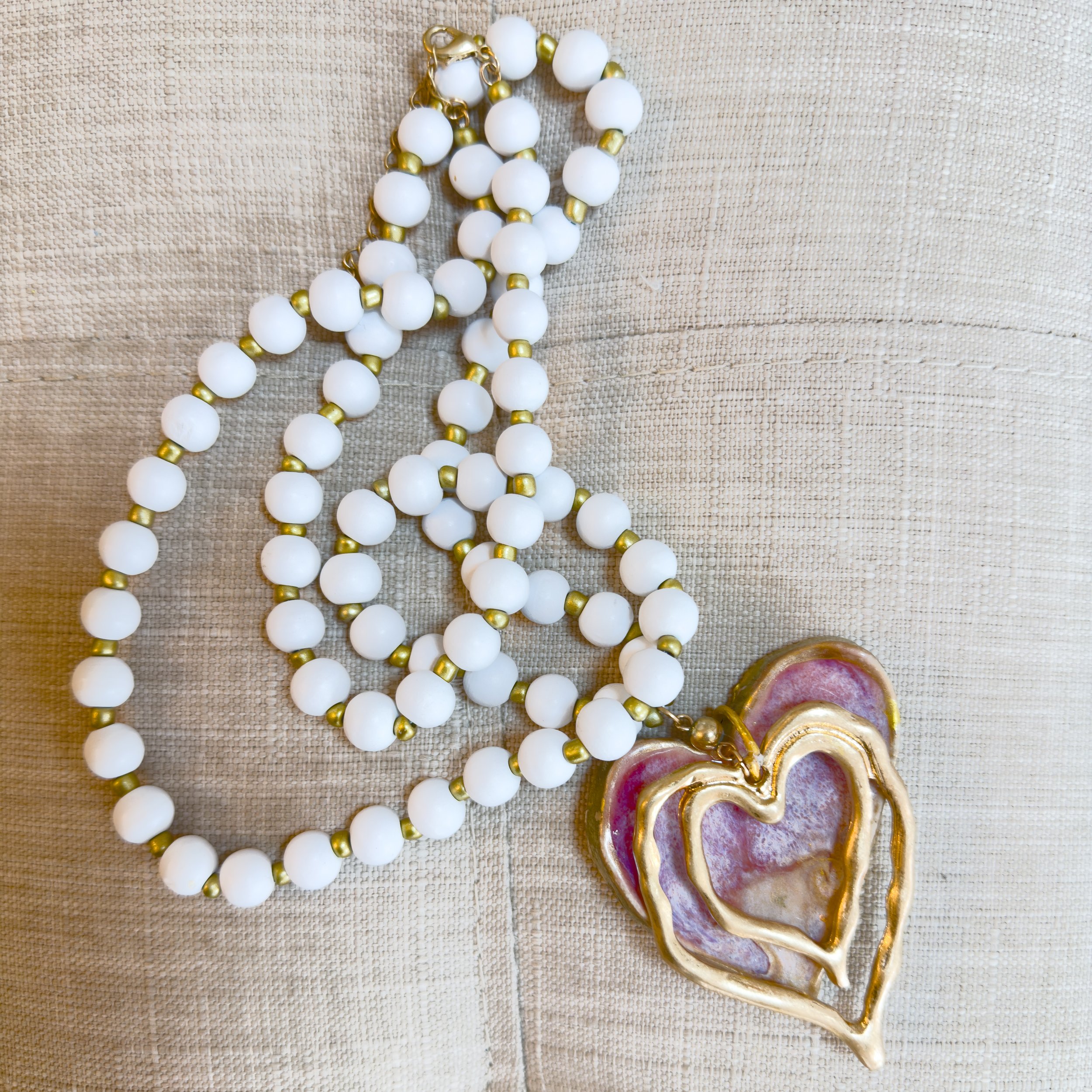 White beaded necklace with a large, decorative, gold-framed heart-shaped pendant on a beige fabric surface.