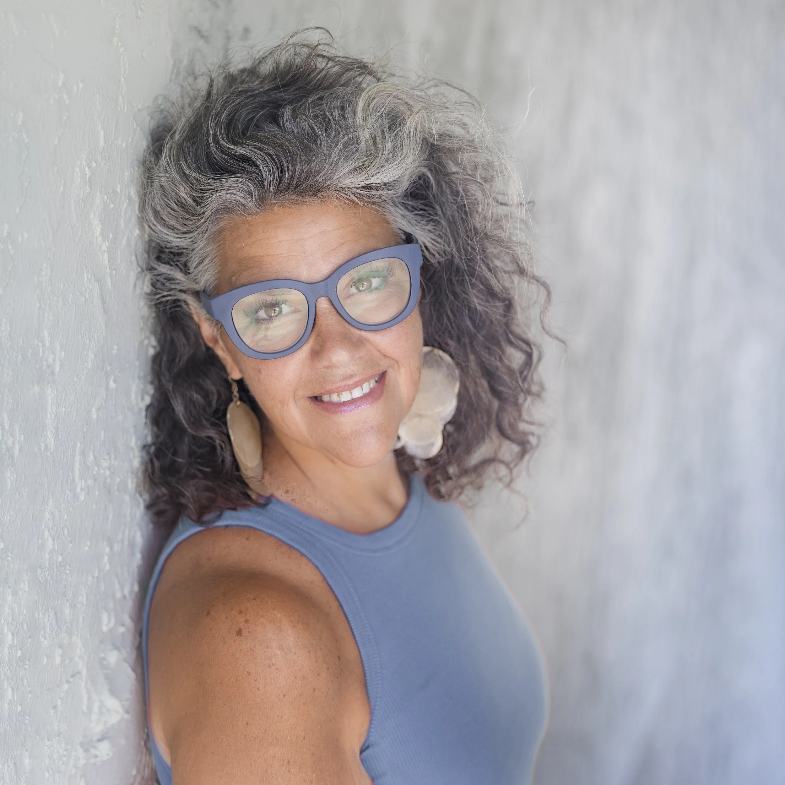 Smiling woman with gray curly hair wearing large beige earrings and blue glasses, dressed in a blue sleeveless top, leaning against a light-colored textured wall.