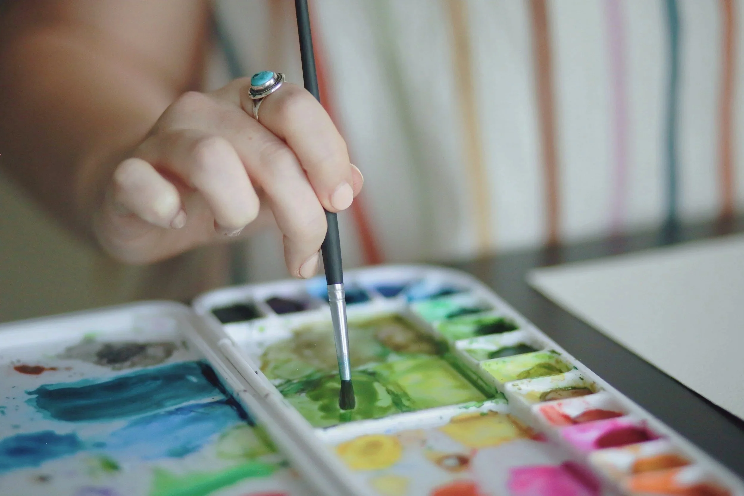 Close-up of a person's hand holding a brush above a watercolor palette with green and blue paints, with a striped curtain in the background.