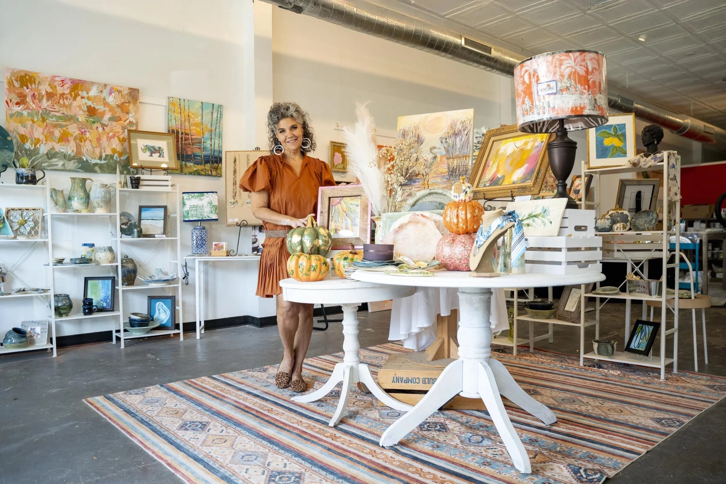 A woman in a rust-colored dress standing behind a table filled with pumpkins, artwork, and decorative items at an art and craft fair. Art paintings and ceramic pieces are displayed on shelves behind her, with a patterned rug on the floor.