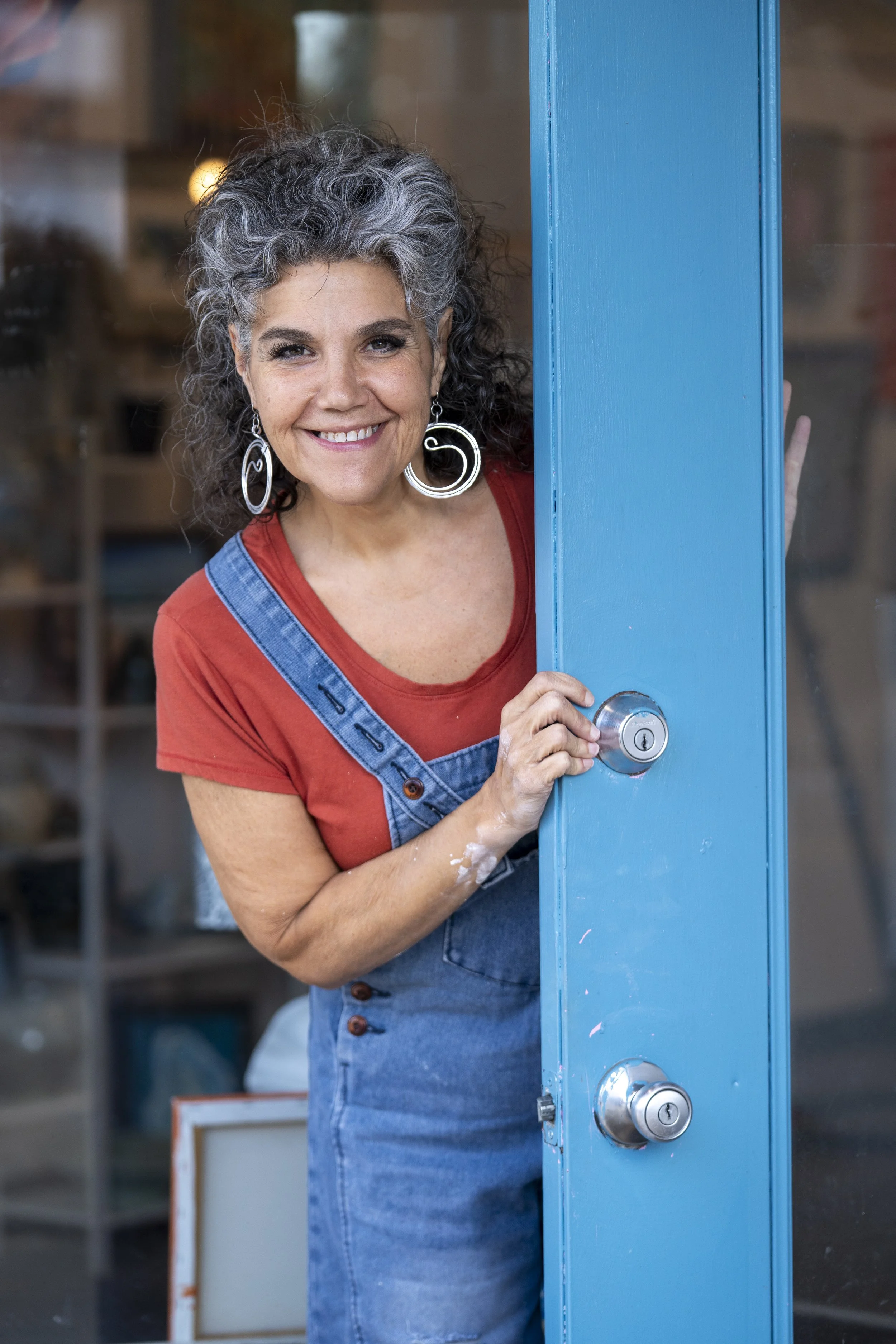 A woman with gray, curly hair smiling and opening a blue door, wearing a red shirt and denim overalls with large hoop earrings.