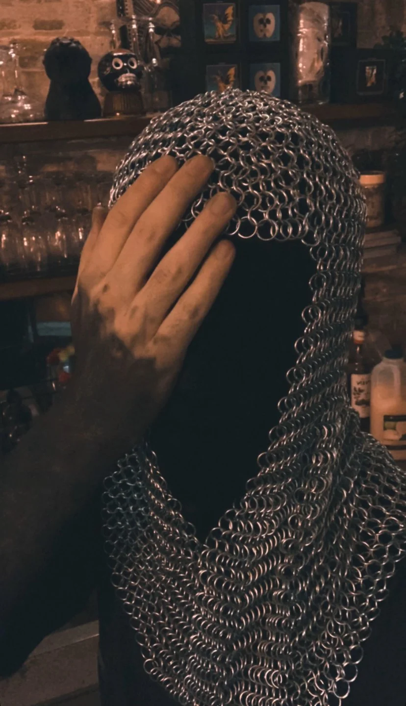 Person wearing a chainmail hood and black clothing, covering their face with their hand in a dimly lit room with shelves of glasses and decorative items.