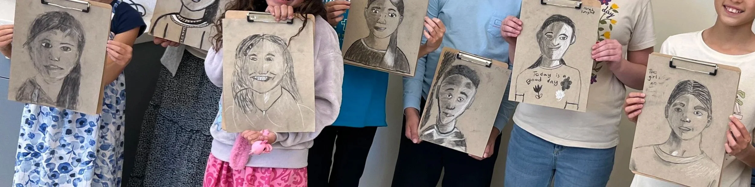 Children holding up portrait sketches of diverse faces on clipboards.
