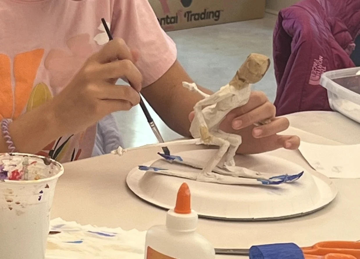 Child painting a papier-mâché sculpture of a dinosaur toy using a paintbrush, construction paper, glue, and art supplies on a white table.