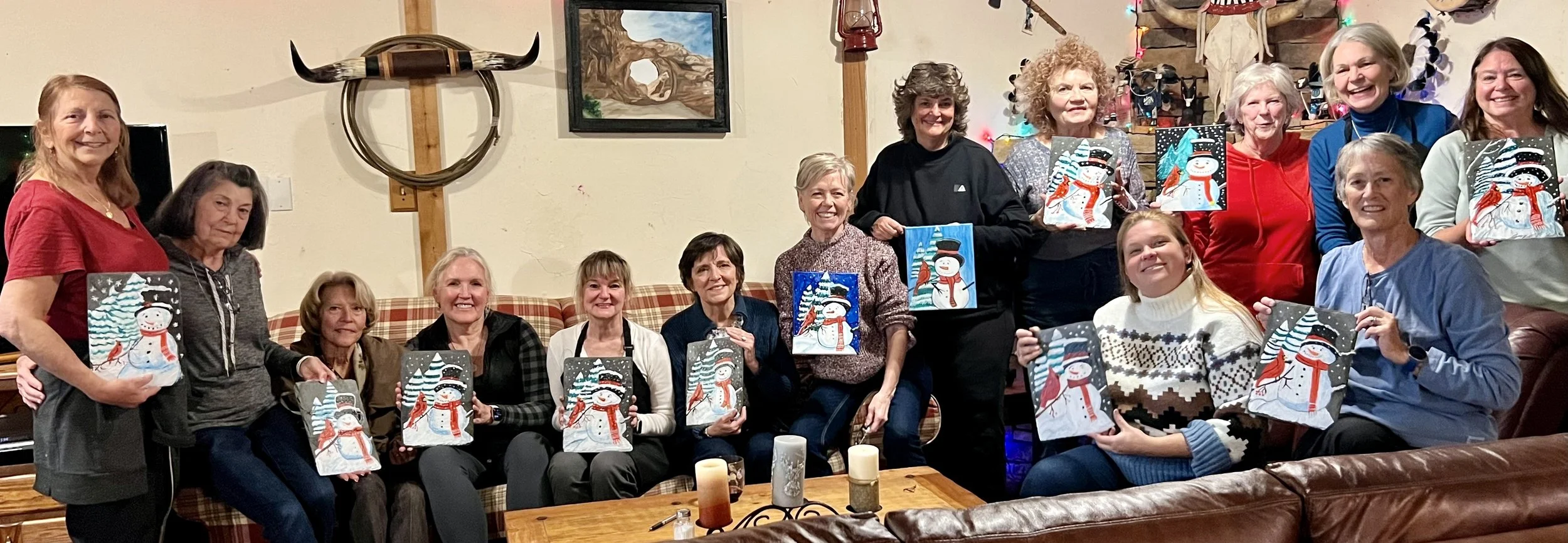 A group of fifteen women gathered in a living room, smiling and holding up snowman-themed holiday gift bags. The room is decorated with Christmas lights, a painting of a natural scene, and decorative items such as candles on a coffee table.