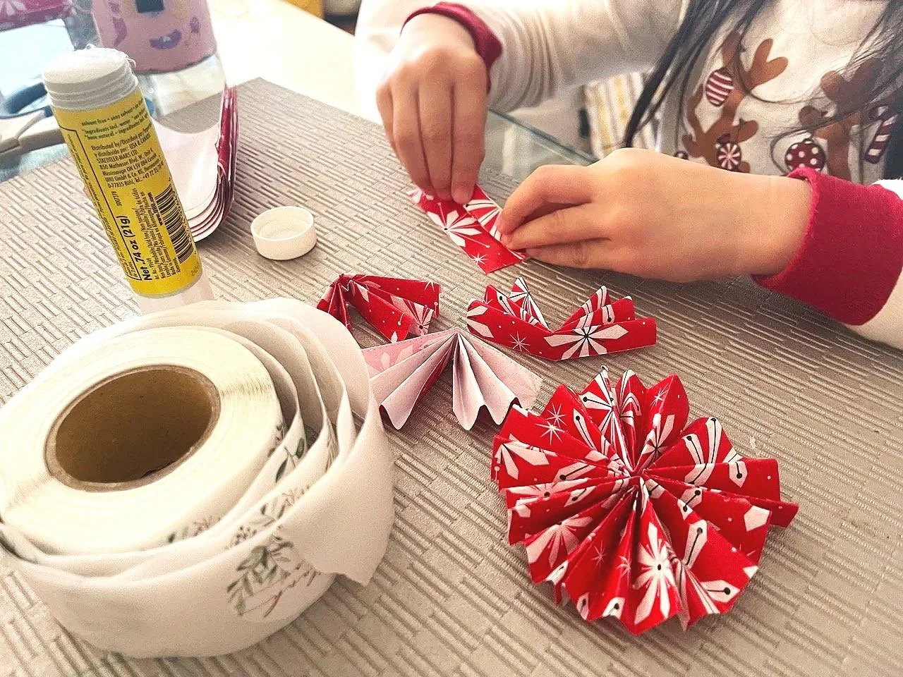 A person making Christmas paper decorations with red patterned paper, surrounded by a roll of white tape, a glue stick, and holiday-themed papers on a table.