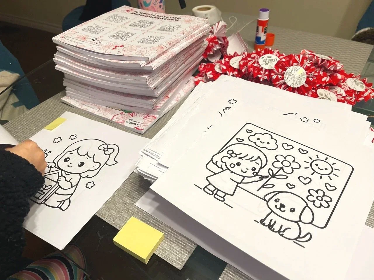Coloring pages with cartoon drawings of a girl holding a gift and a girl presenting a flower with a dog, on a table with stacks of similar pages, holiday-themed decorations, glue, and red patterned paper.