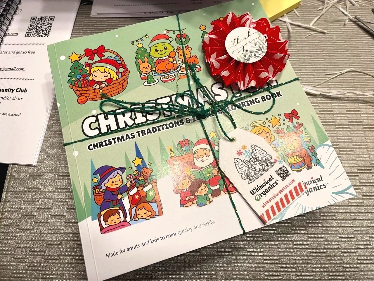 Coloring book titled 'Christmas Traditions & Holiday Coloring Book' with Christmas-themed illustrations and a red origami flower on top, tied with green string, on a table.