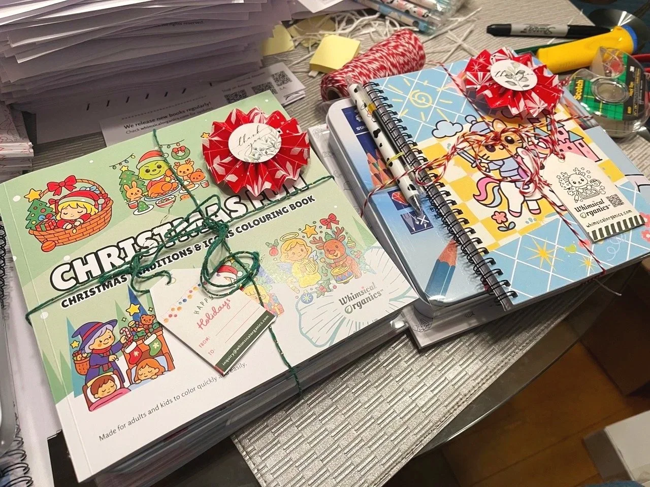 Stack of Christmas coloring books tied with ribbon, decorated with red and white ribbon rosettes, sitting on a cluttered desk with office supplies and papers.