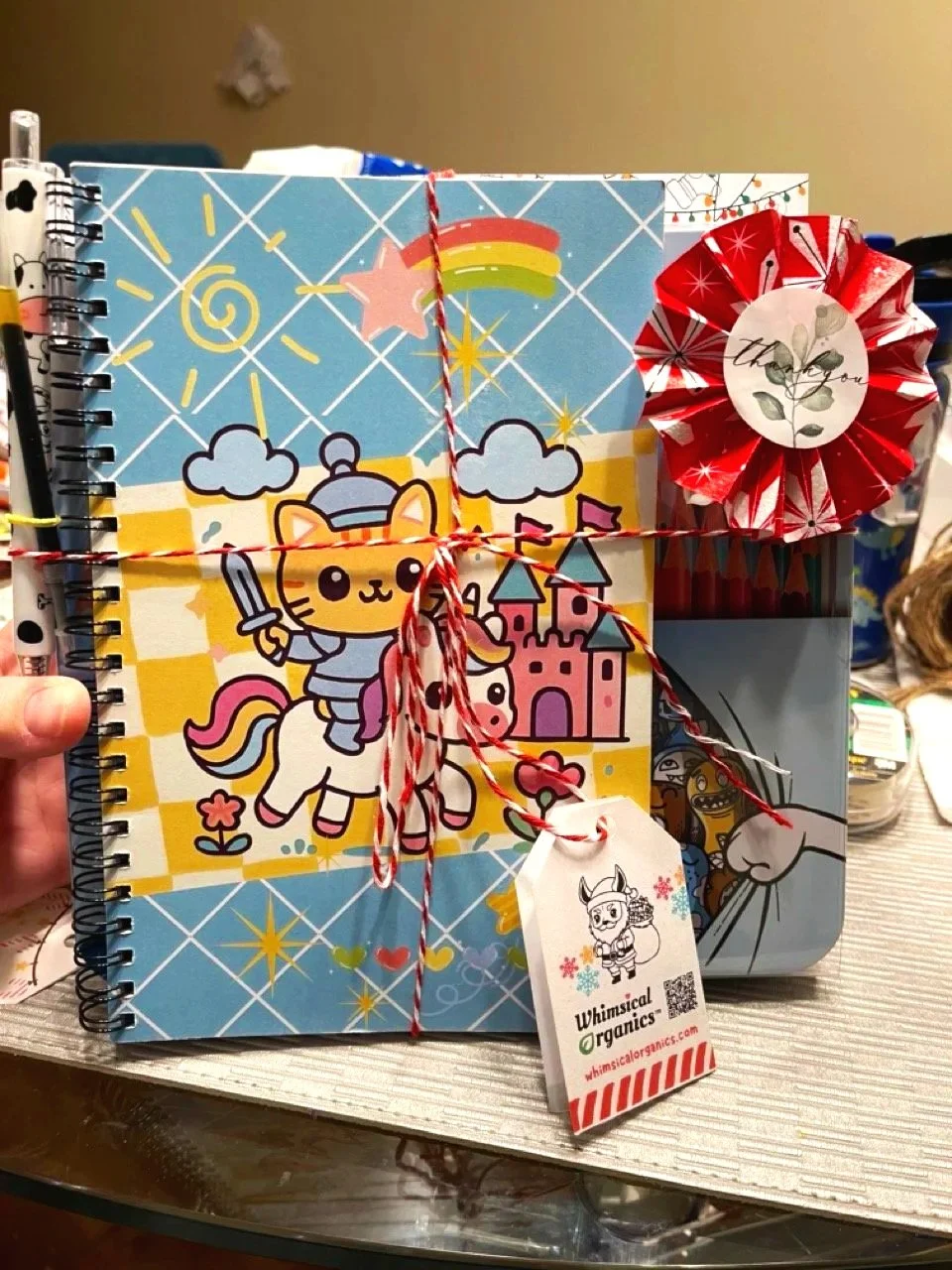 A colorful notebook with a cute cat knight drawing, decorated with a red and white ribbon, a red paper rosette, and a tag from Whimsical Organics. The notebook is set against a cluttered background.