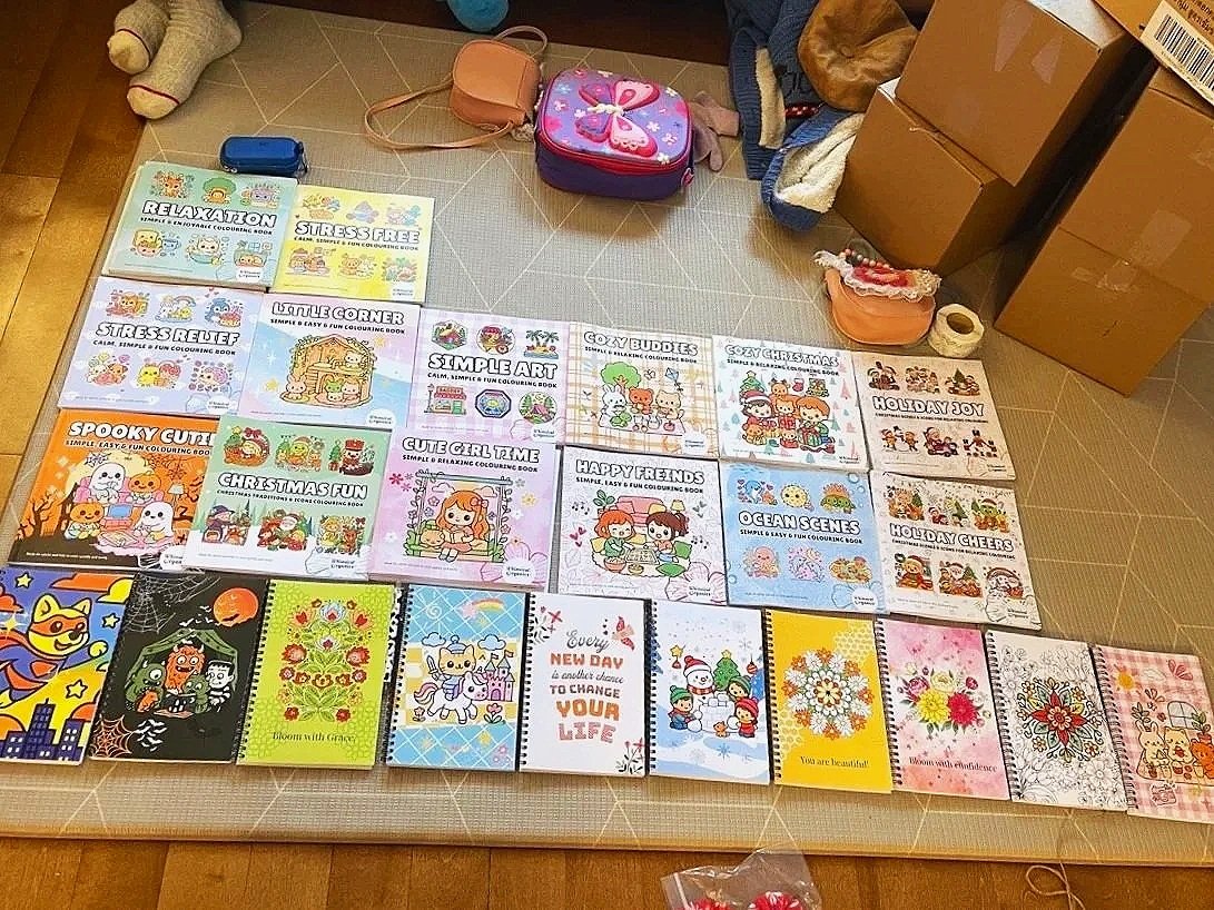 A collection of coloring books and notebooks arranged on the floor, with boxes, bags, and miscellaneous items in the background.