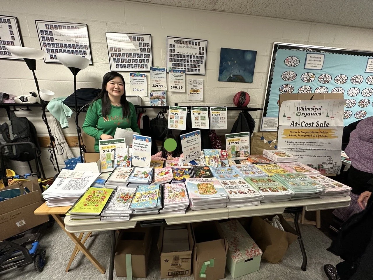 A woman in a green shirt standing behind a table full of holiday-themed coloring books and activity pages at a fundraising sale event in a school gymnasium, with posters and signs displaying prices and discounts on the wall behind her.