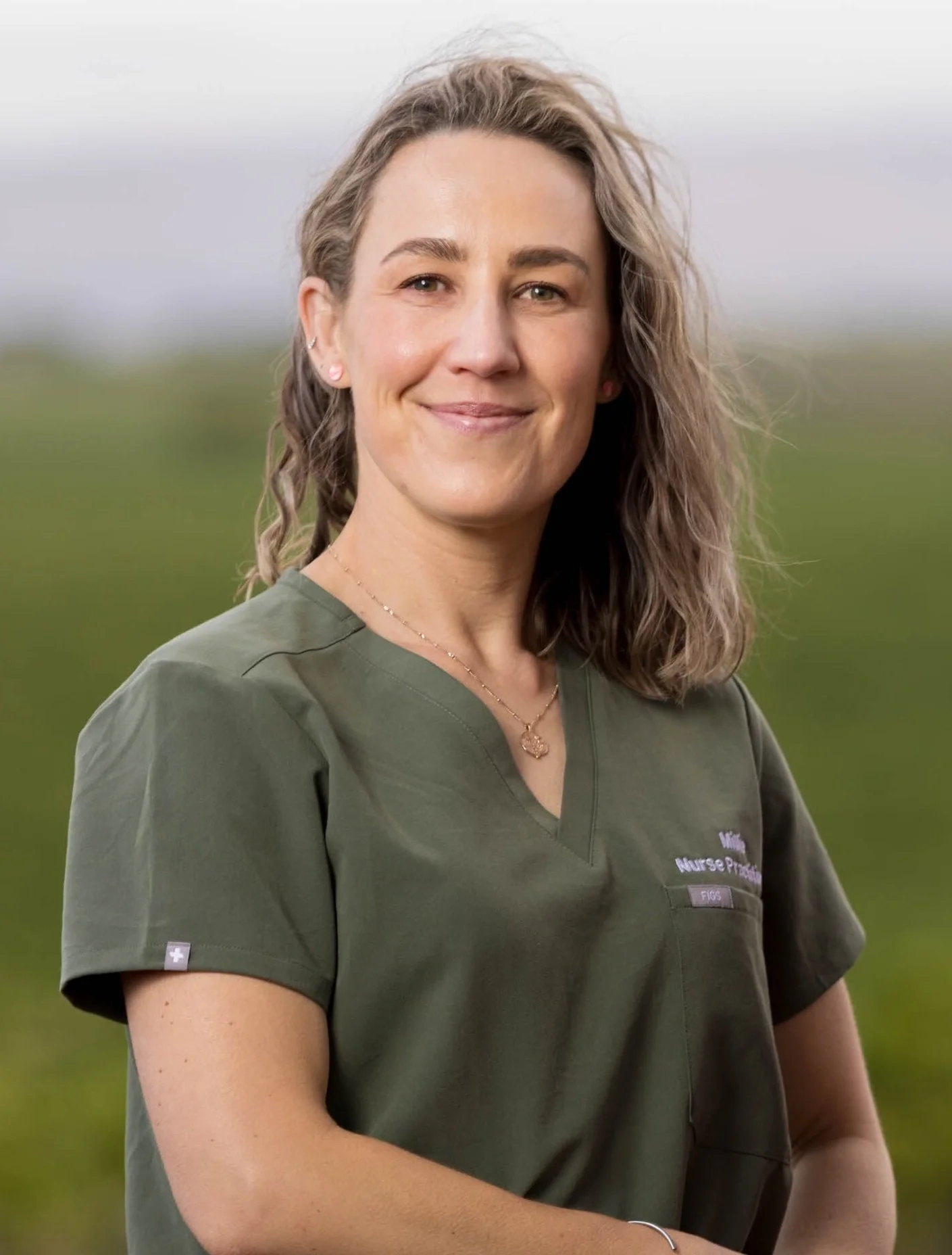 A smiling female nurse standing outdoors in a green field, wearing a green medical uniform with a badge.