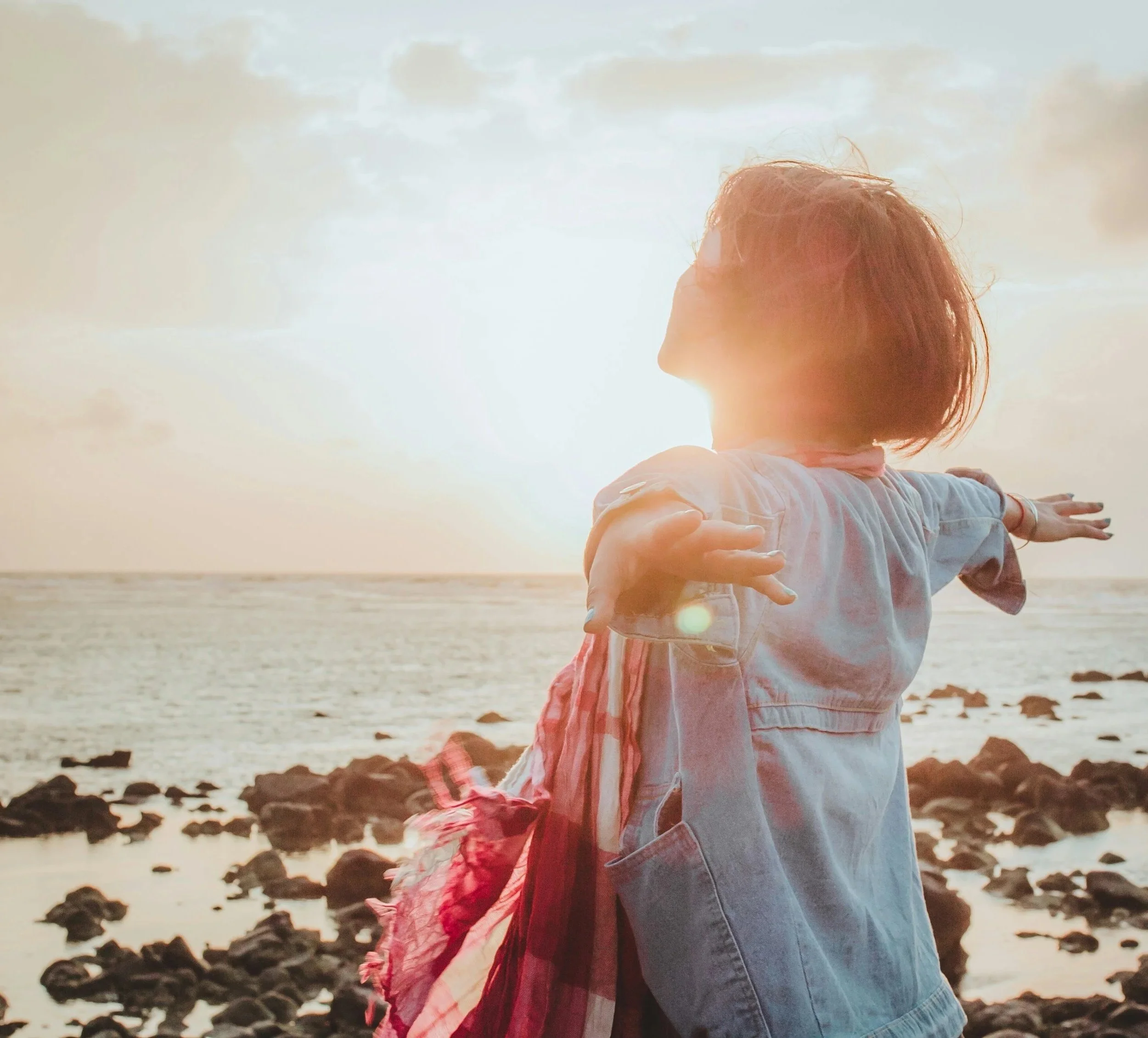 A young girl with short hair wearing a denim shirt and a plaid jacket, arms outstretched, enjoying a sunset at the rocky beach.