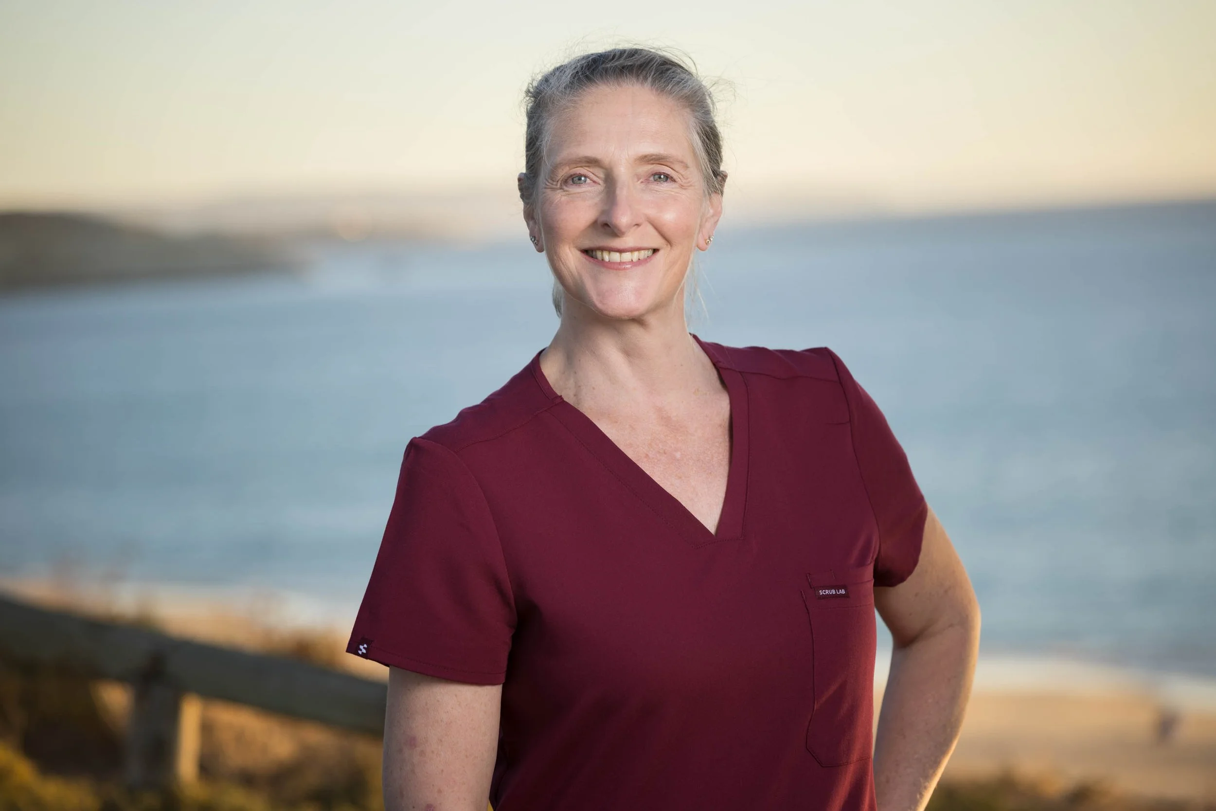 A woman wearing a maroon scrub top, standing outdoors near a beach with water and sky in the background, smiling, with gray hair tied back.