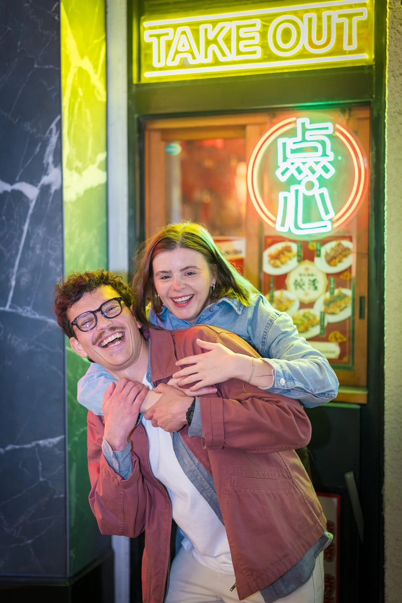 Two people, a man with glasses and a woman, smiling and laughing outside a Chinese takeout restaurant, illuminated by neon signs.