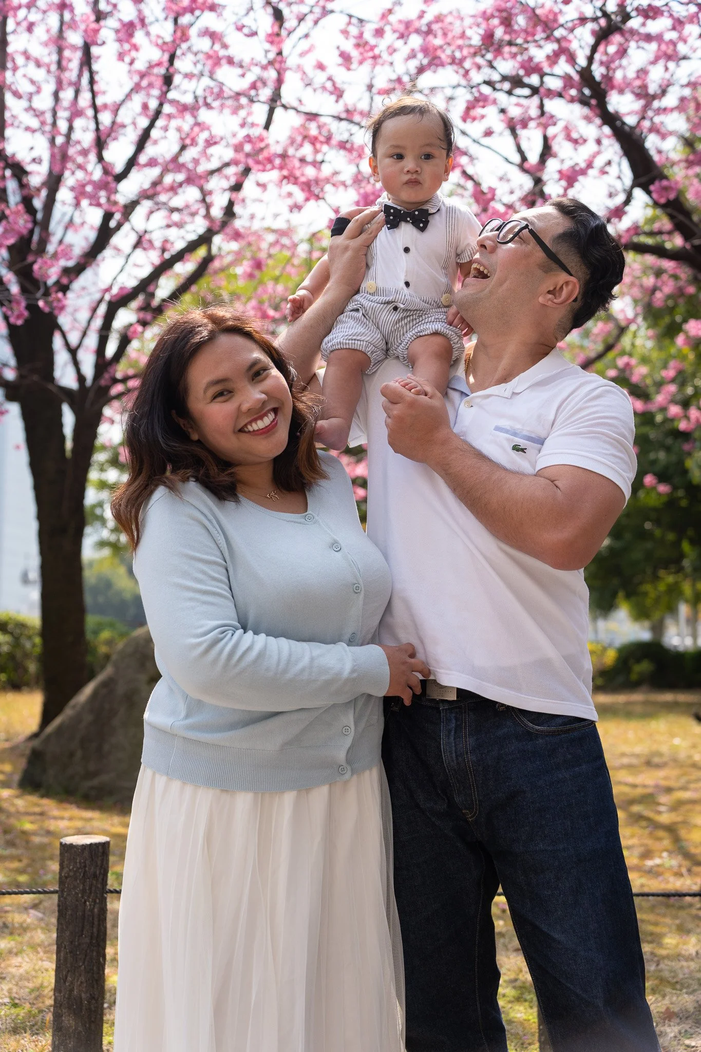 A family of three enjoying time outdoors under blossoming pink cherry trees in a park. The woman is smiling, the man is holding a young child wearing a white shirt with a bow tie, and they are surrounded by greenery and rocks.