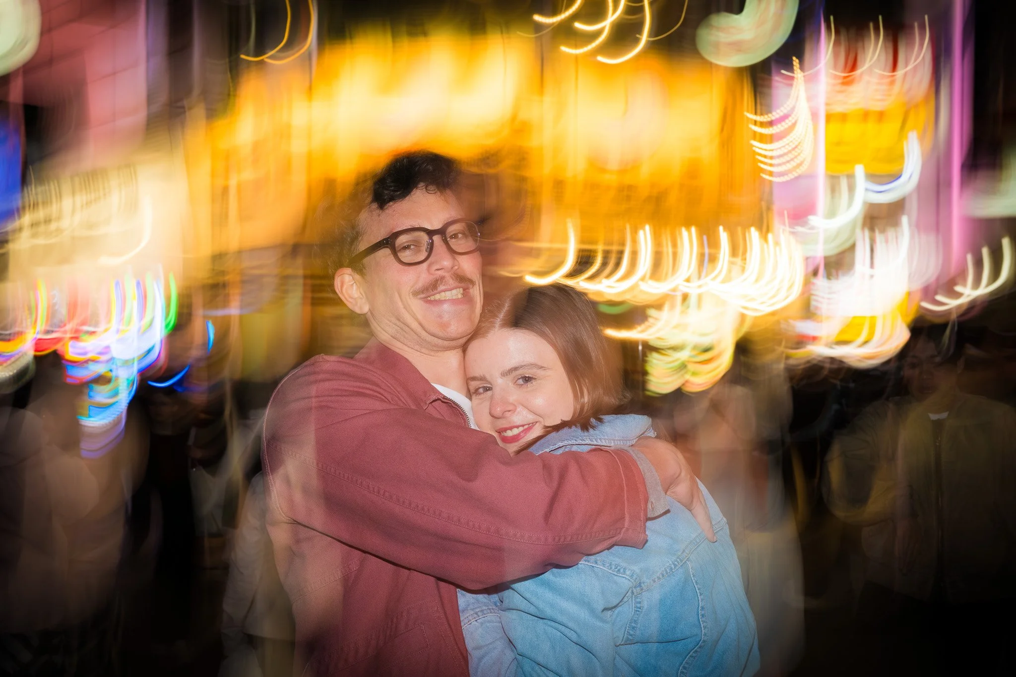 A man with glasses and a woman with dark hair embracing and smiling in a setting with colorful, blurred neon lights in the background.