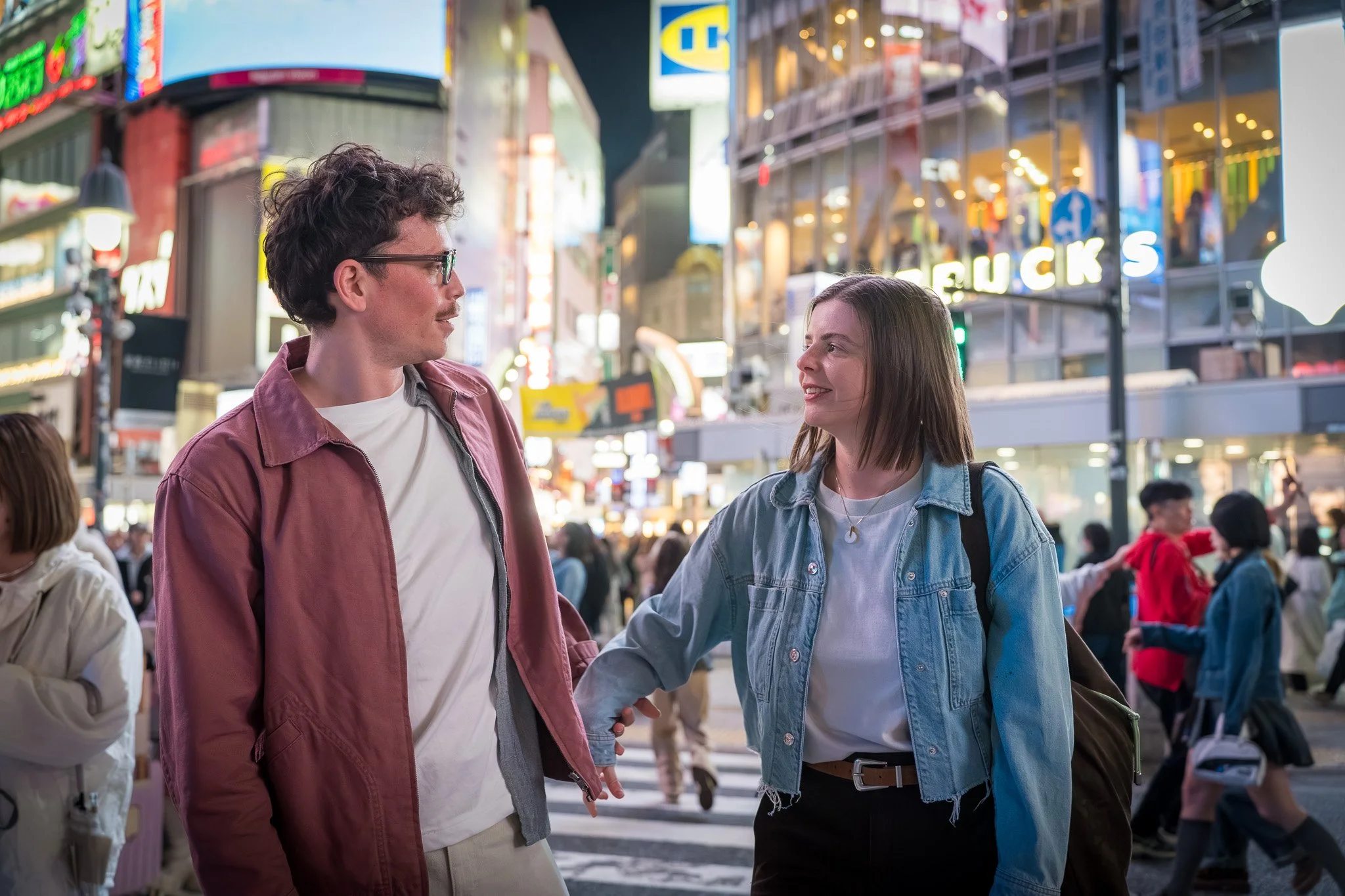A young man and woman are talking and holding hands on a busy city street at night, illuminated by bright neon signs and storefront lights.