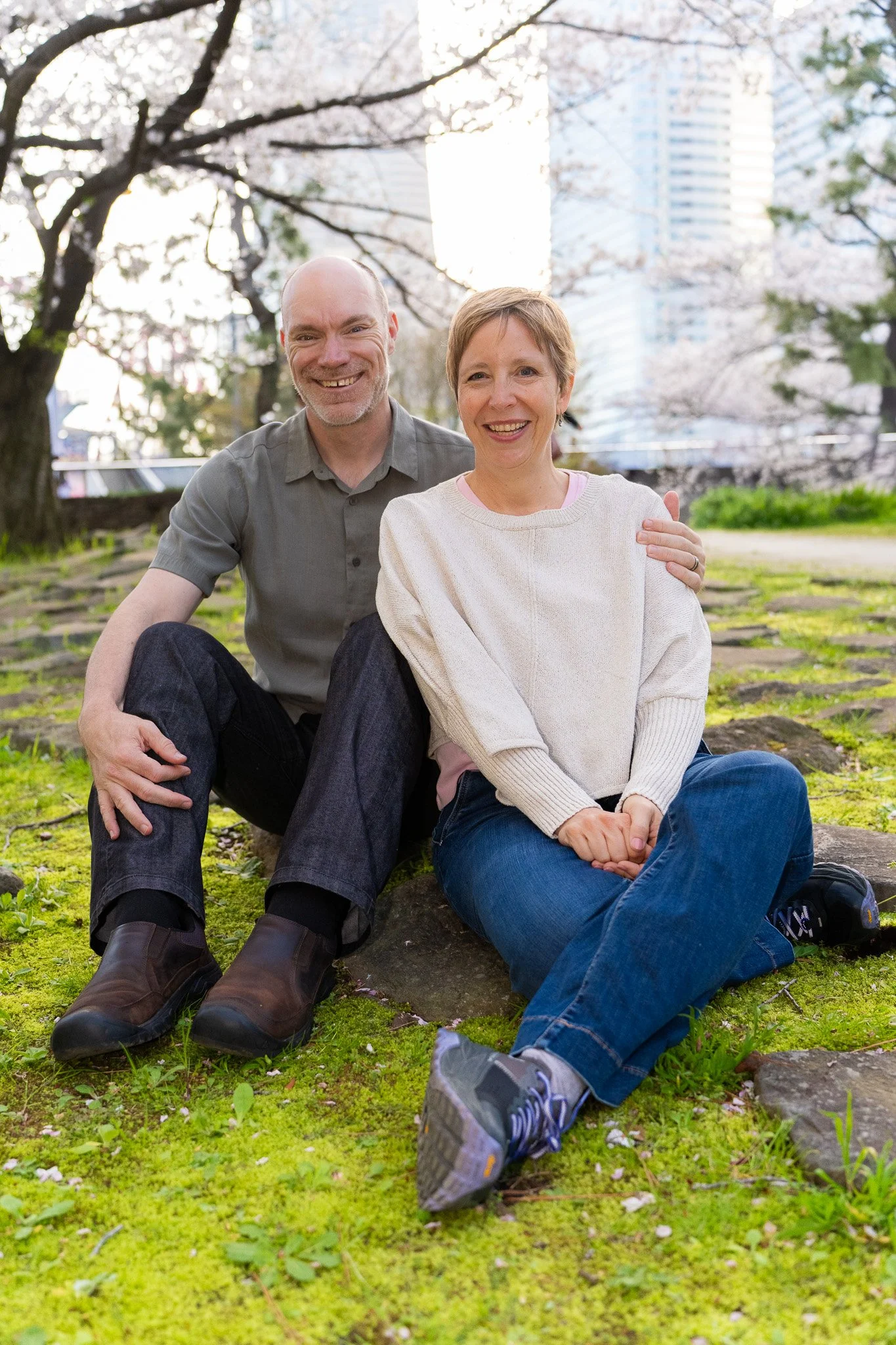 A happy couple sitting on the grass in a park during spring, with blooming cherry blossom trees and tall city buildings in the background.