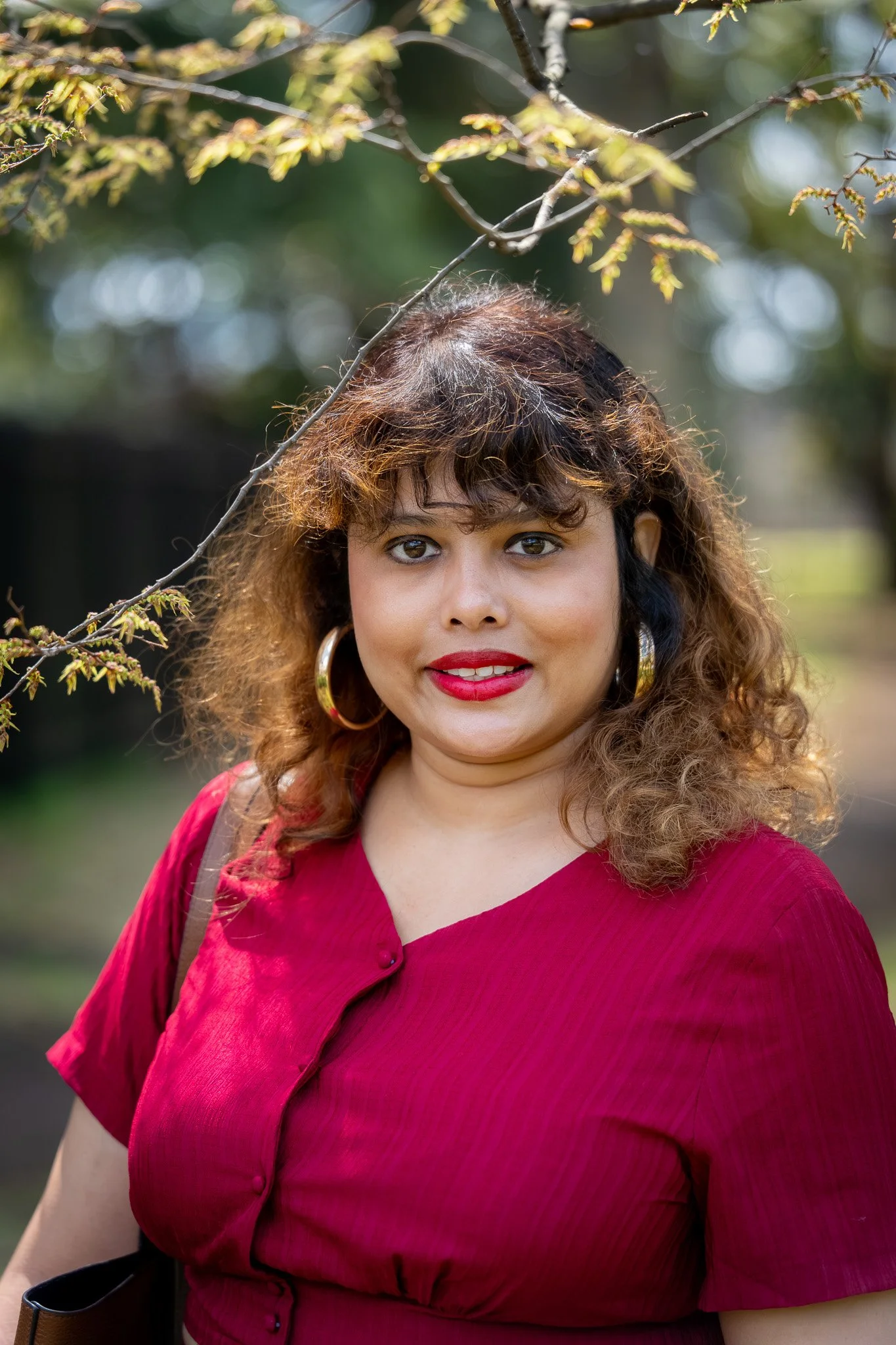 A woman with curly hair, wearing a red top and gold hoop earrings, standing outdoors with trees and foliage in the background.