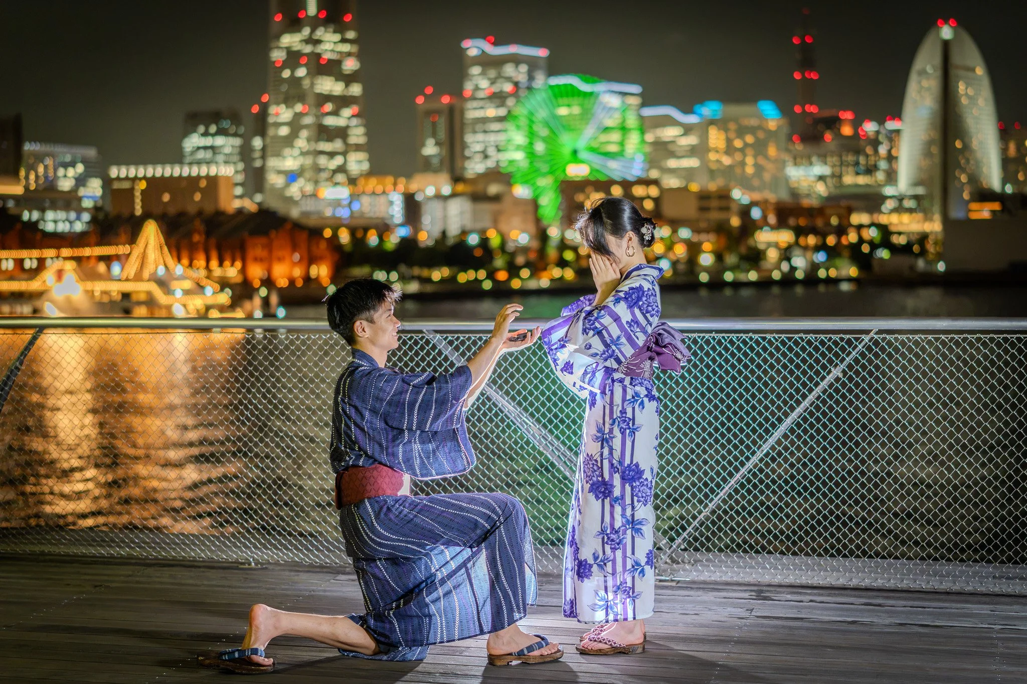 Kneeling man proposing to woman on a deck at night with city skyline and colorful lights in the background.