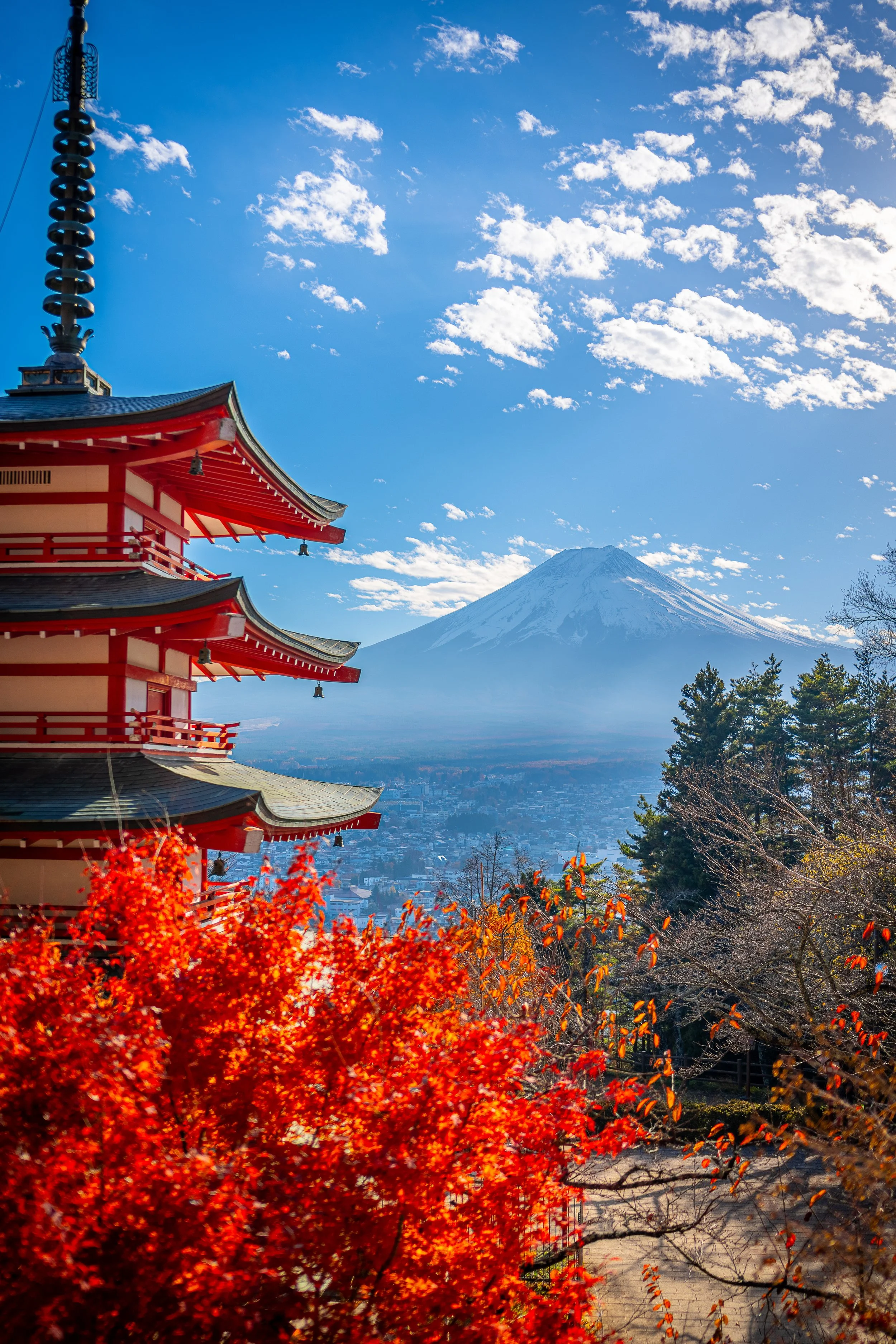 A view of Mount Fuji with a traditional Japanese pagoda in the foreground, surrounded by vibrant autumn trees, under a bright blue sky with scattered clouds.