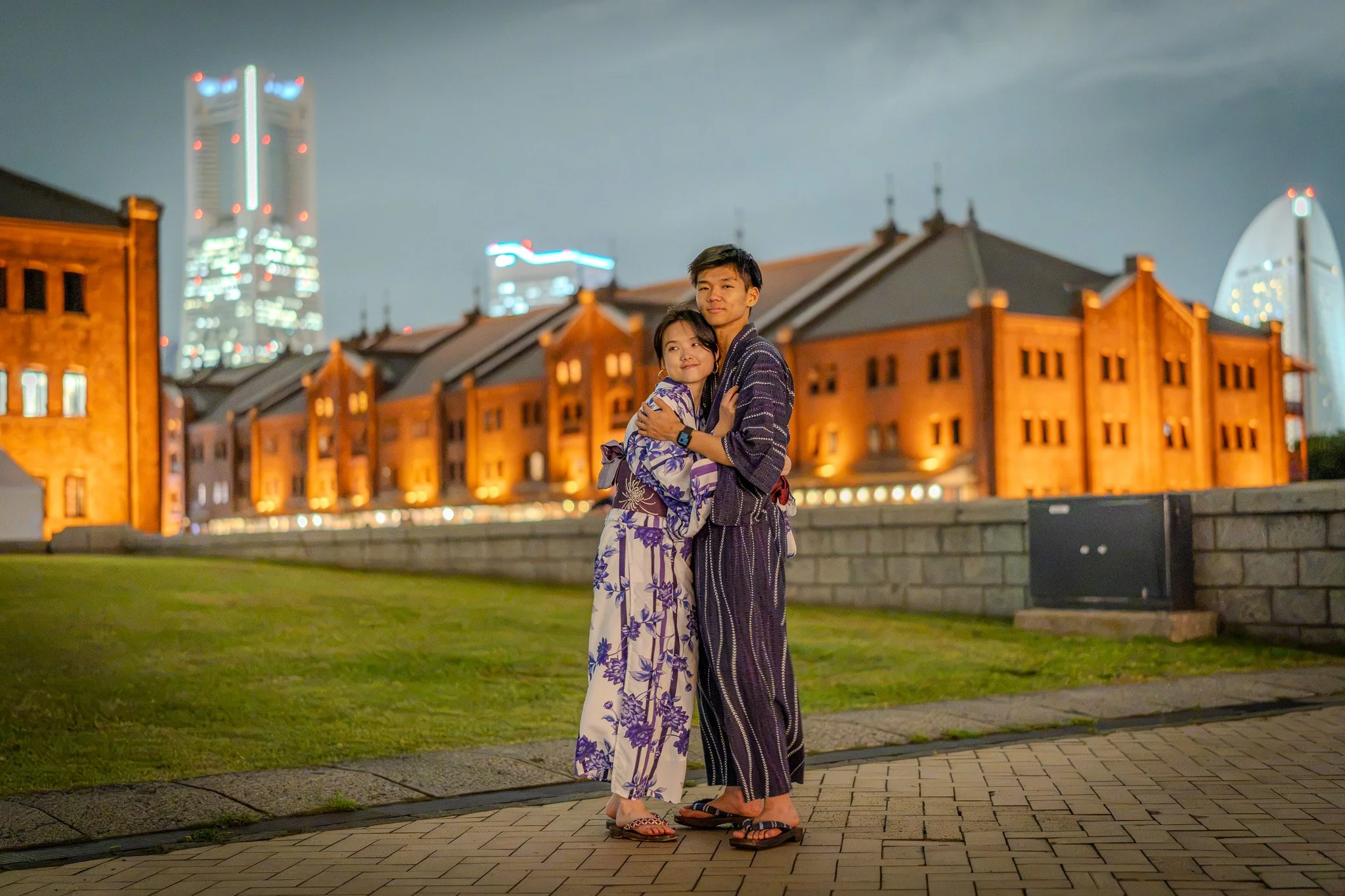 Two people standing close together at night in front of illuminated buildings, embracing and wearing traditional Japanese yukata.
