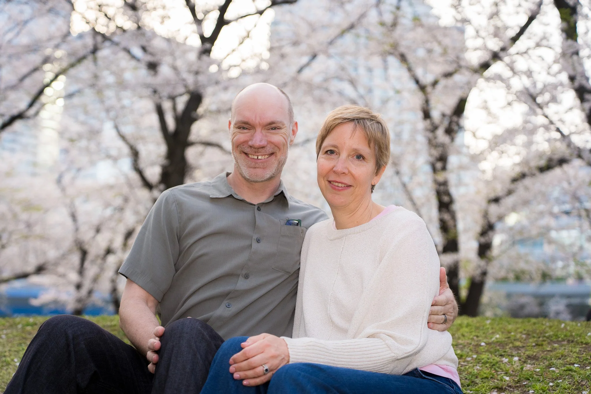 A happy middle-aged couple sitting together on grass in a park with blooming cherry blossom trees in the background.