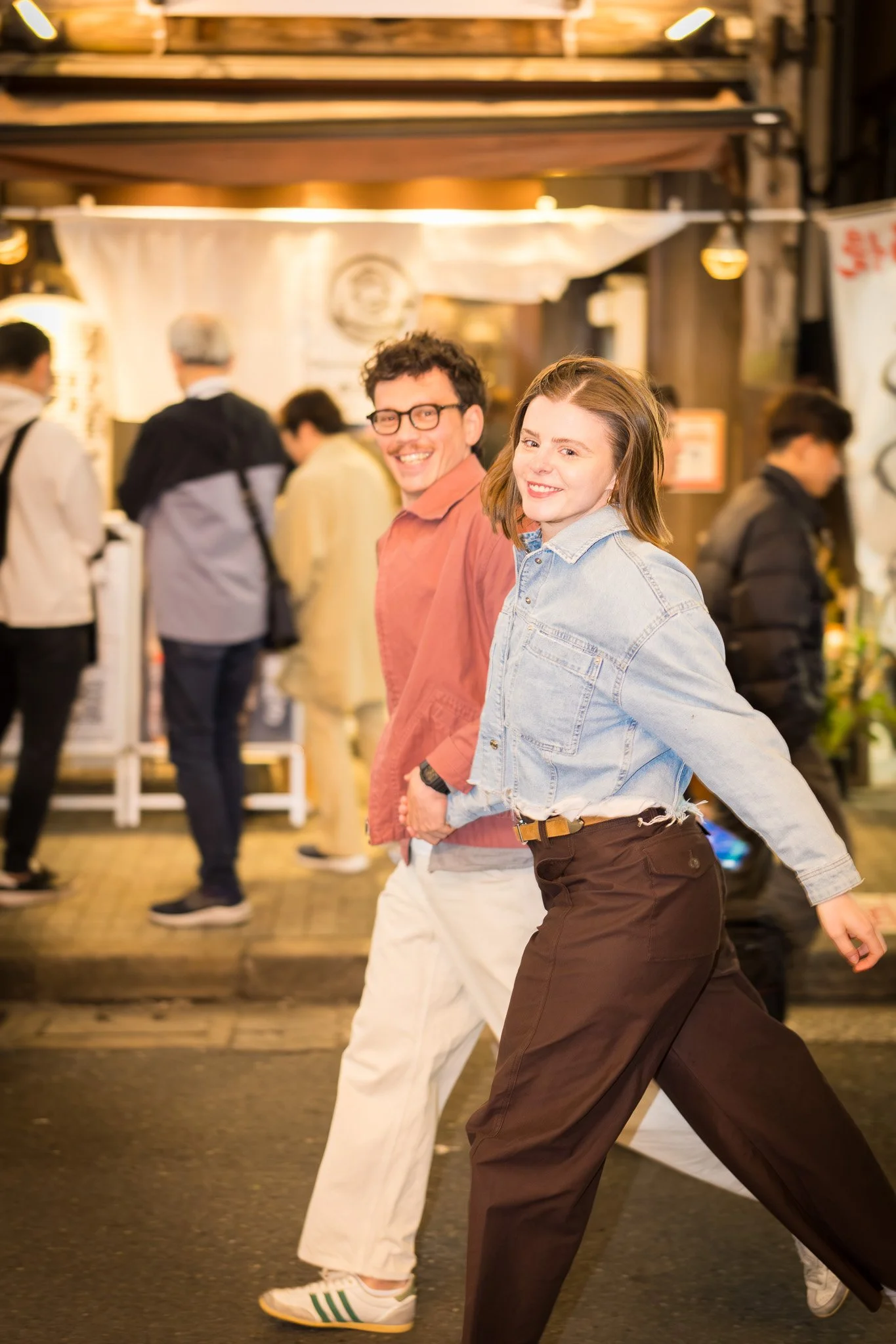 Two smiling people, a man in a salmon shirt and white pants and a woman in a denim jacket and dark pants, walking in a busy street at night with people in the background.