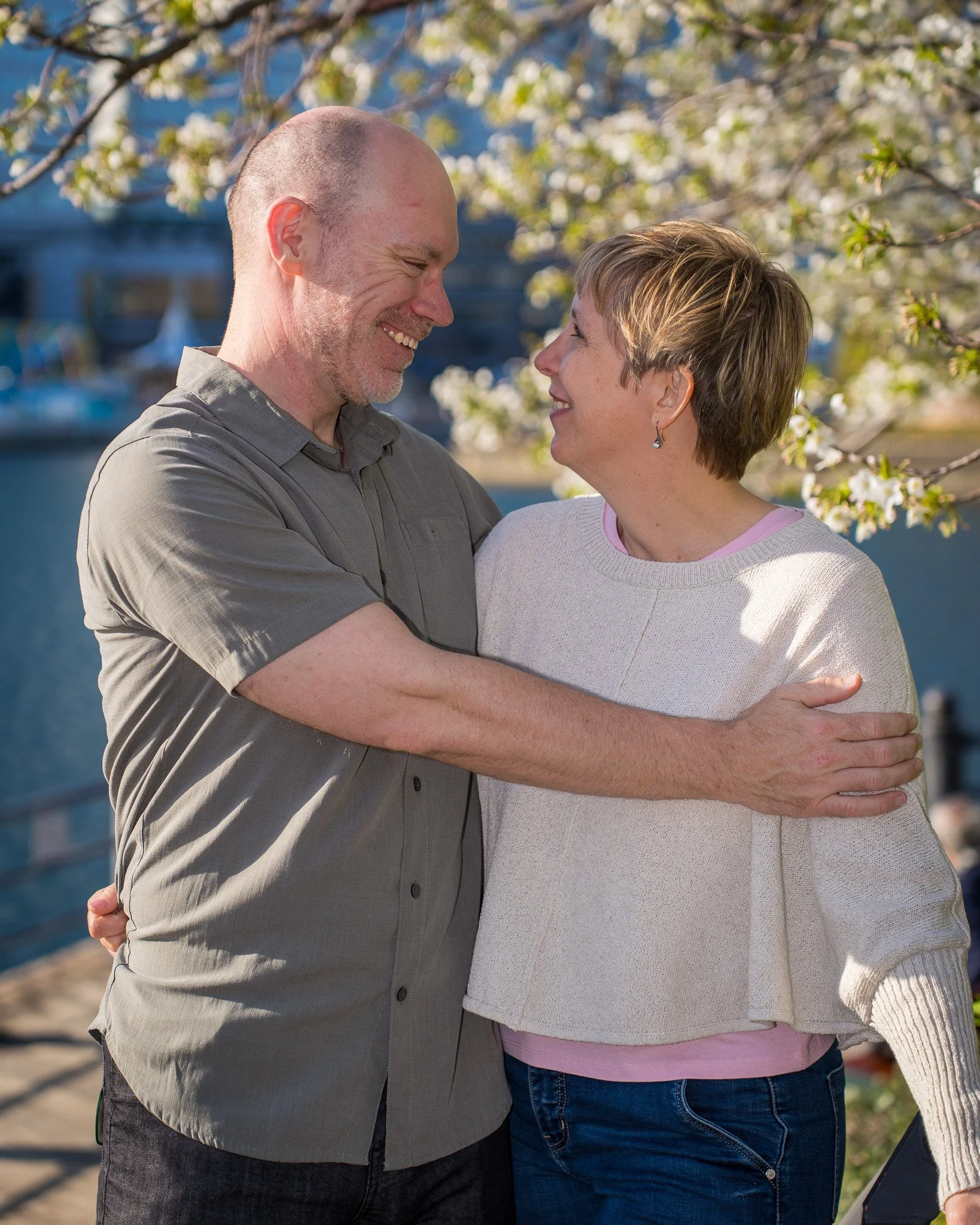 A couple embracing outdoors near a body of water with trees and boats, smiling at each other.