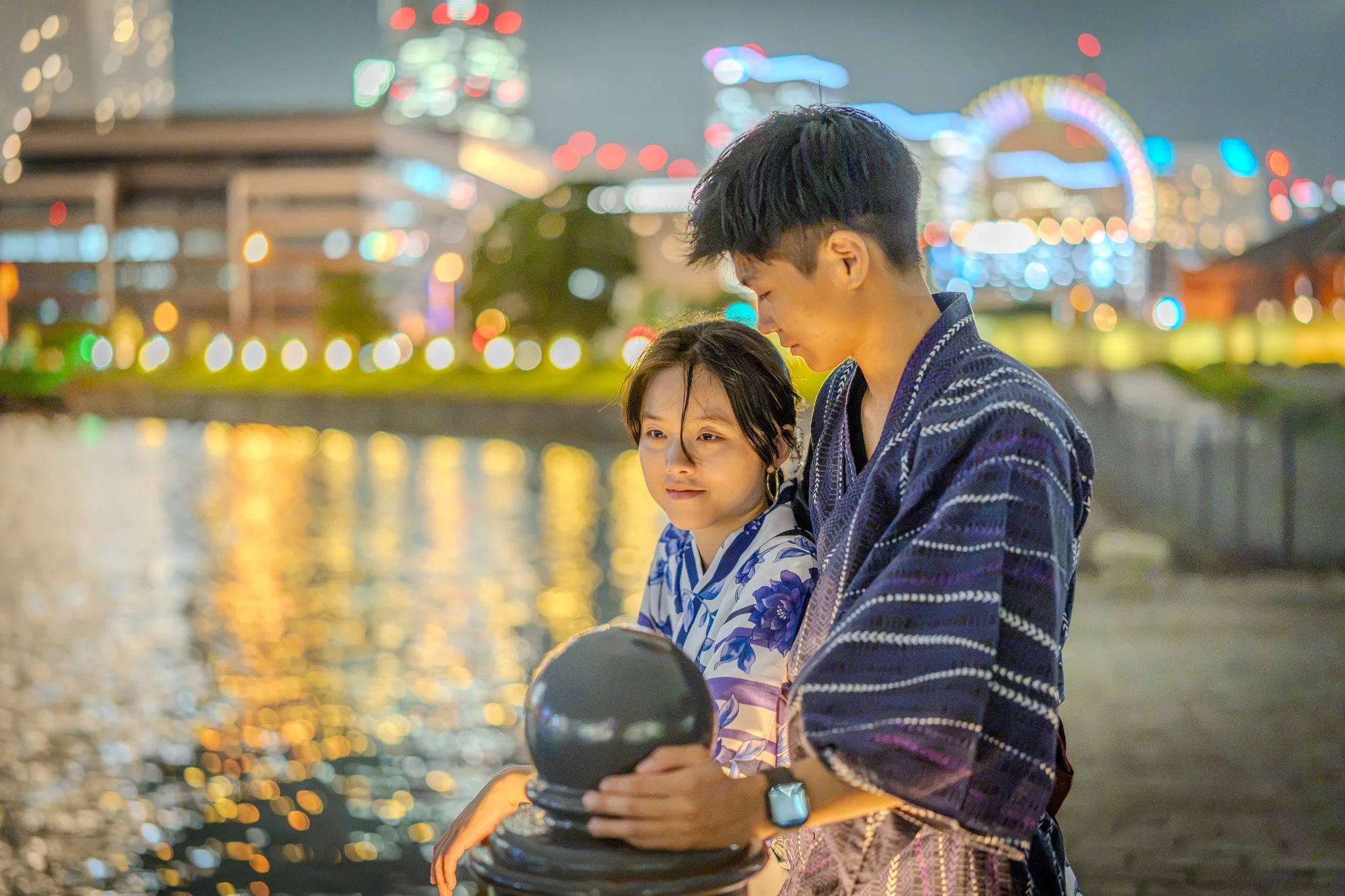 A young man and a girl wearing yukatas sitting by a river at night, with colorful lights and a Ferris wheel in the background.
